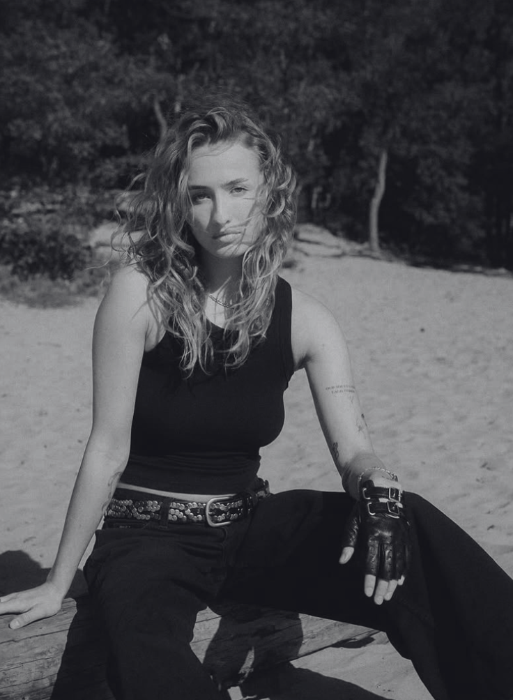 A young woman with curly hair sitting outdoors on a wooden surface, wearing a sleeveless top, black pants, a belt, and a fingerless glove, with a background of trees and a sandy area.