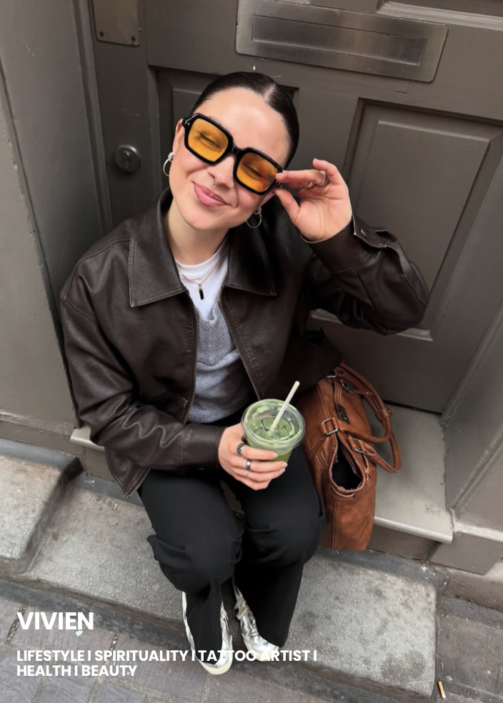 A woman with dark hair, wearing sunglasses with orange lenses, a brown leather jacket, a gray sweater, and black pants, is sitting on a sidewalk next to a gray door. She is smiling and holding a drink with a straw. There is a brown leather bag beside her.