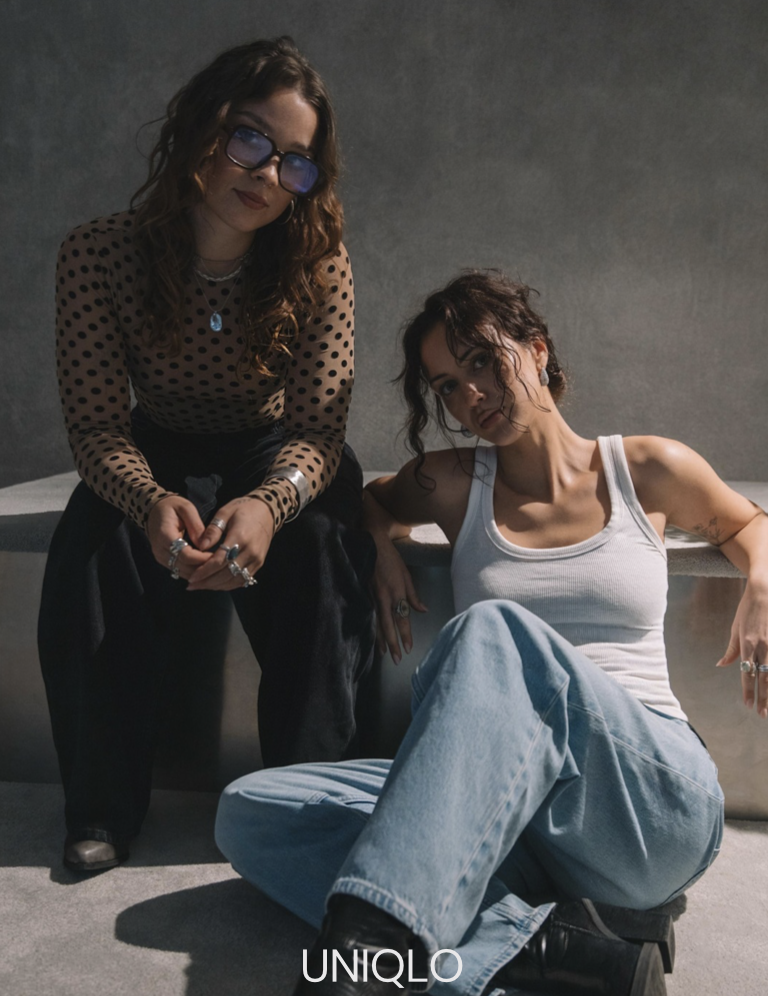 Two women posing together in a modern, minimalist setting. The woman on the left is standing, wearing large glasses, a polka dot top, and jewelry. The woman on the right is sitting on the floor, wearing a white tank top and loose jeans, with curly hair.