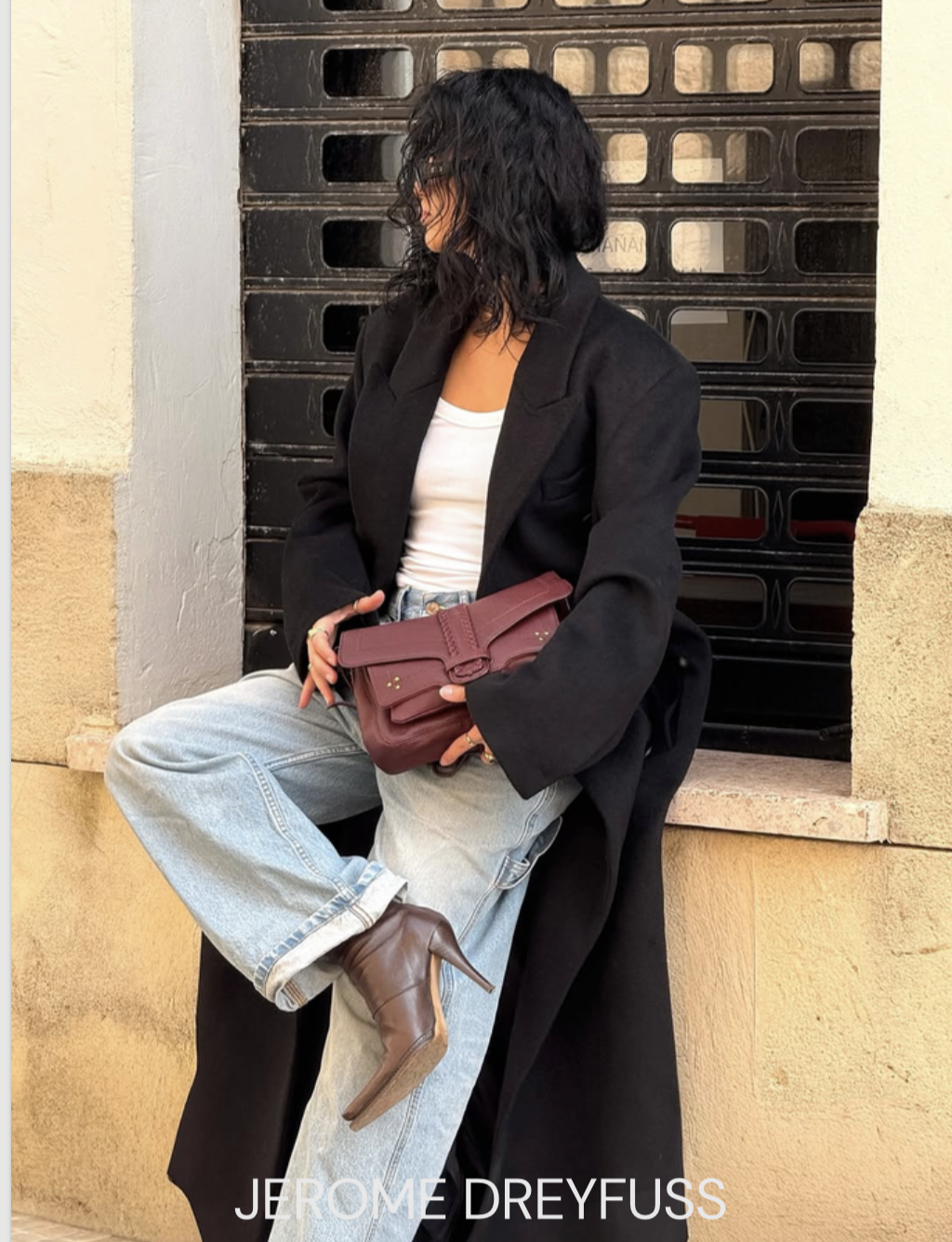 A woman with curly black hair wearing a black coat, white top, light blue jeans, and brown high-heeled boots, sitting on a ledge with one leg crossed over the other, holding a maroon purse, in front of a black metal gate and beige wall.