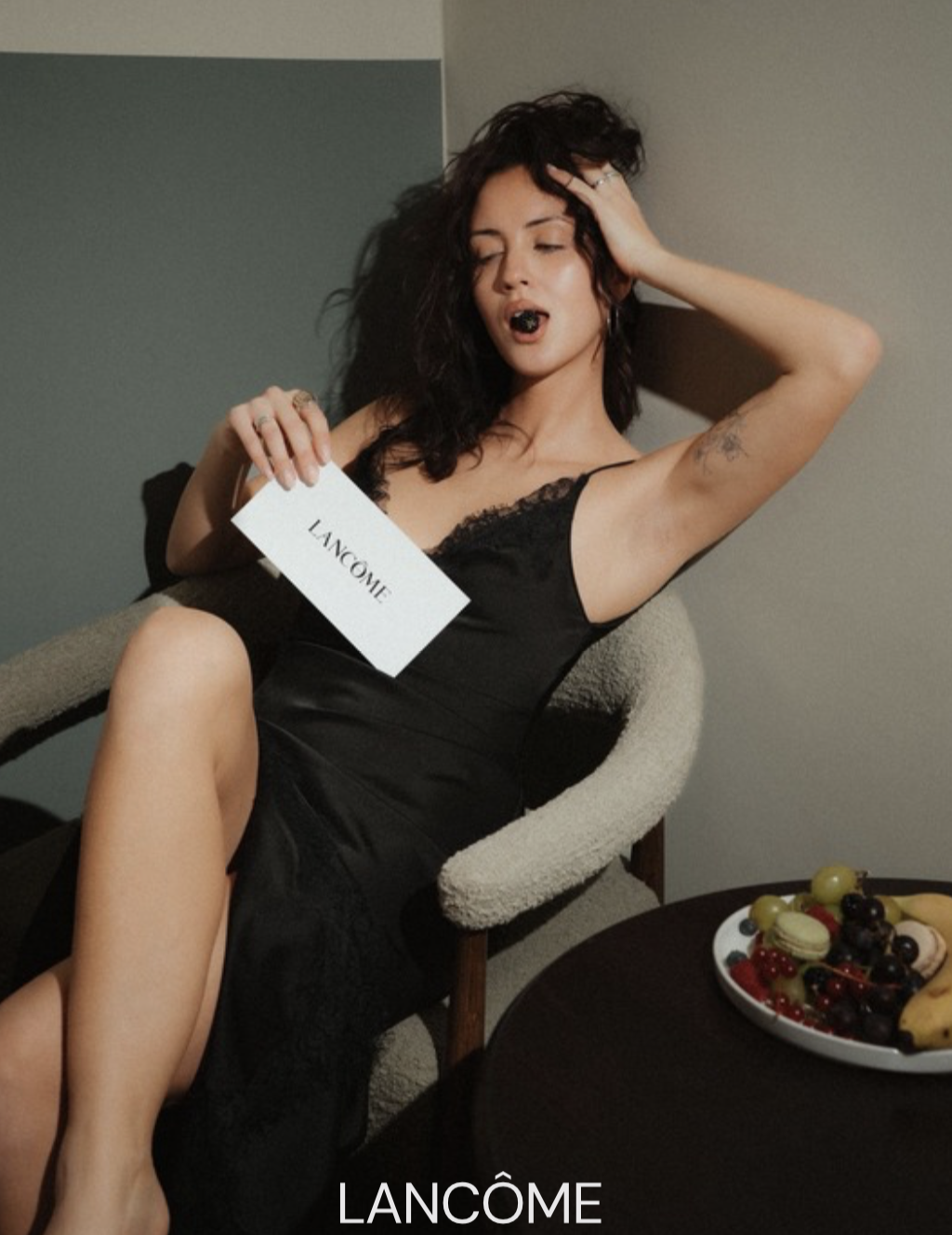 A woman with curly dark hair in a black lace and satin dress, sitting on a beige armchair, holding a Lancôme advertisement card, with a lipstick or makeup stain on her lips, and a plate of fruit on the table in front of her.