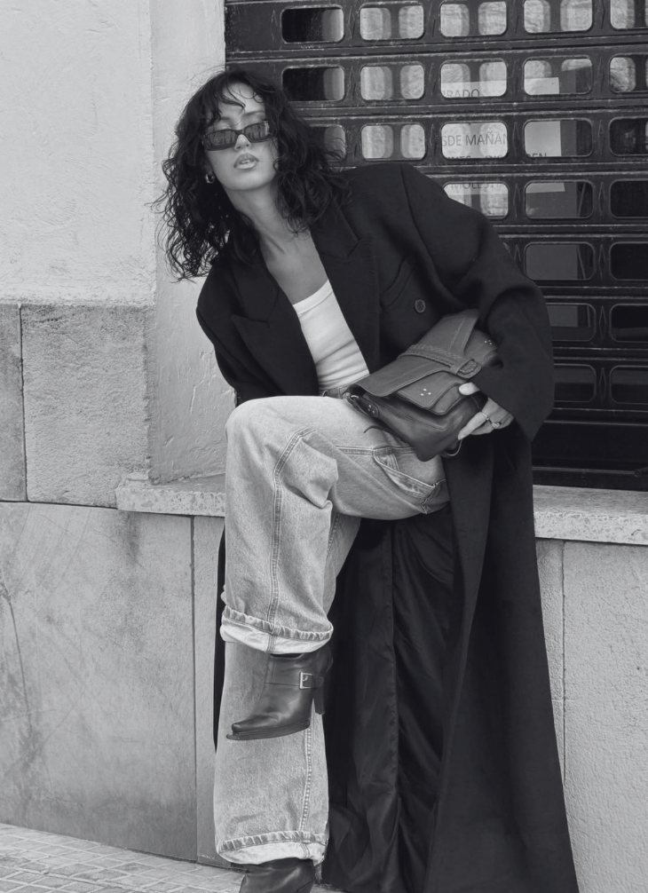 A woman with curly hair wearing sunglasses, a long coat, a white shirt, baggy jeans, and black boots, sitting against a wall near a metal gate.