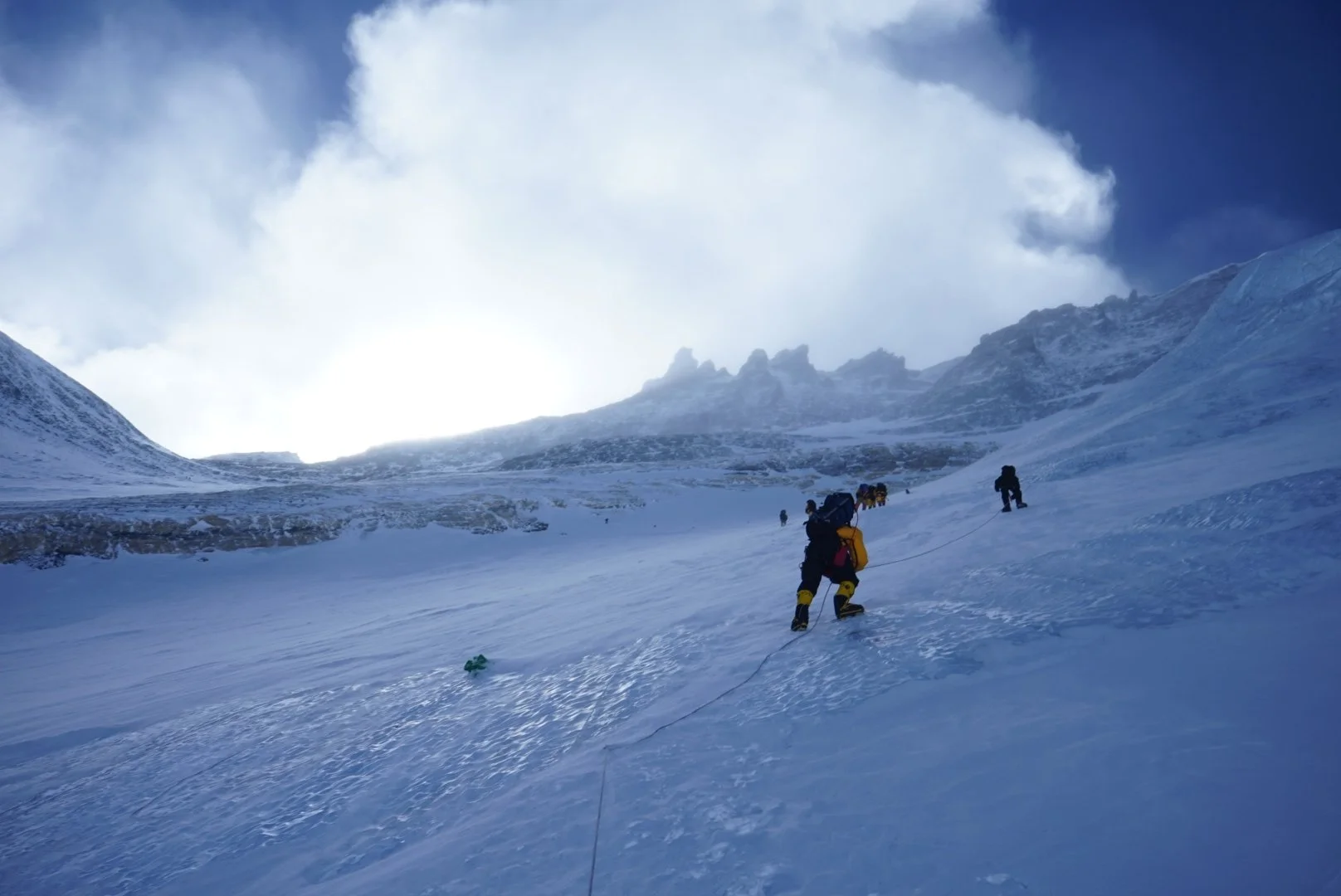 Mountaineers ascending a snowy mountain slope, with cloudy sky and rugged peaks in the background.
