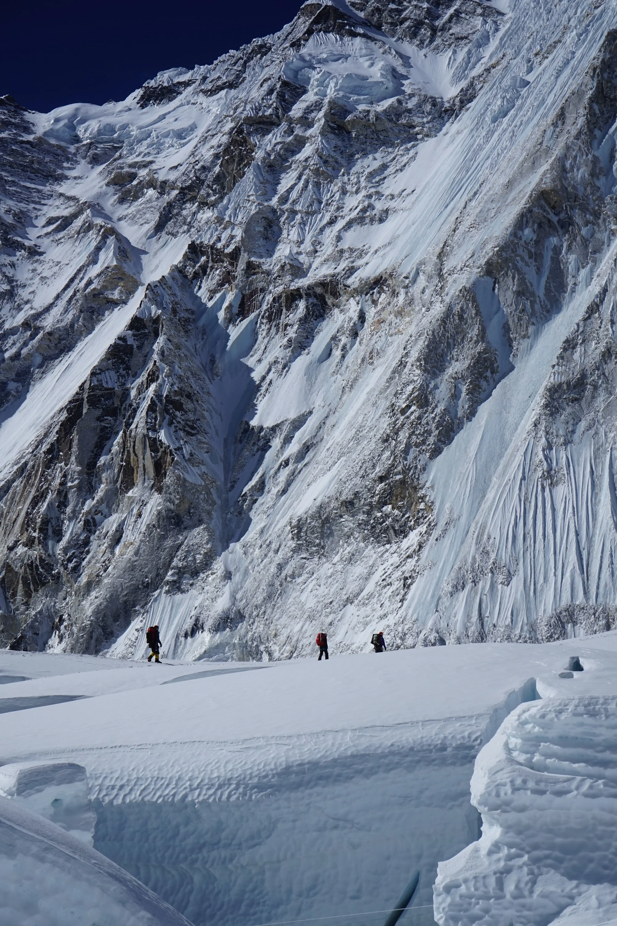 Three mountaineers in colorful gear trekking through a snow-covered mountain landscape with steep, jagged peaks and icy slopes.