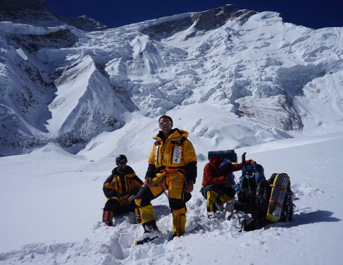 Three mountaineers in yellow and red jackets resting in snow with large snow-covered mountain in the background.