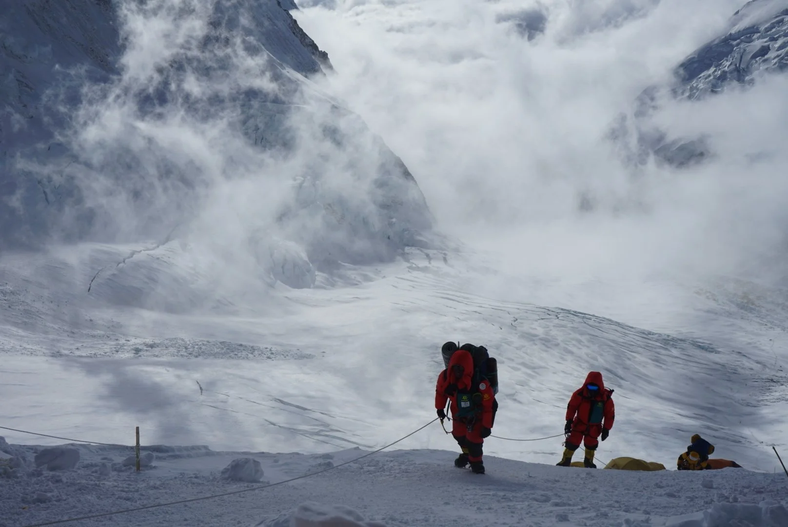 Two mountaineers in red heavy-duty winter gear climbing on snow and ice in a high-altitude mountain environment, with a large crevasse or avalanche area in the background, surrounded by snow-covered peaks and clouds.