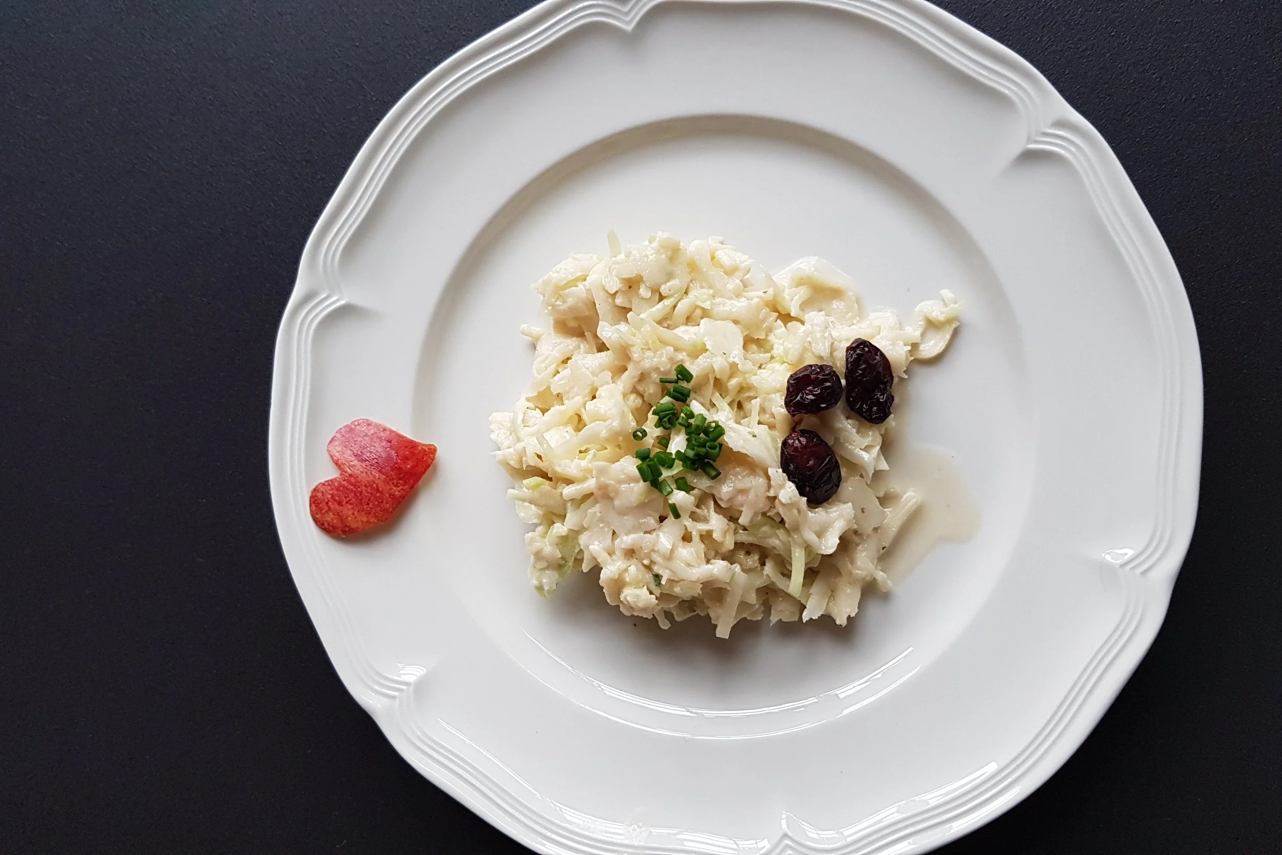 Ein weißer Teller mit Nudelsalat, garniert mit Schnittlauch und getrockneten Preiselbeeren, auf einem schwarzen Hintergrund.