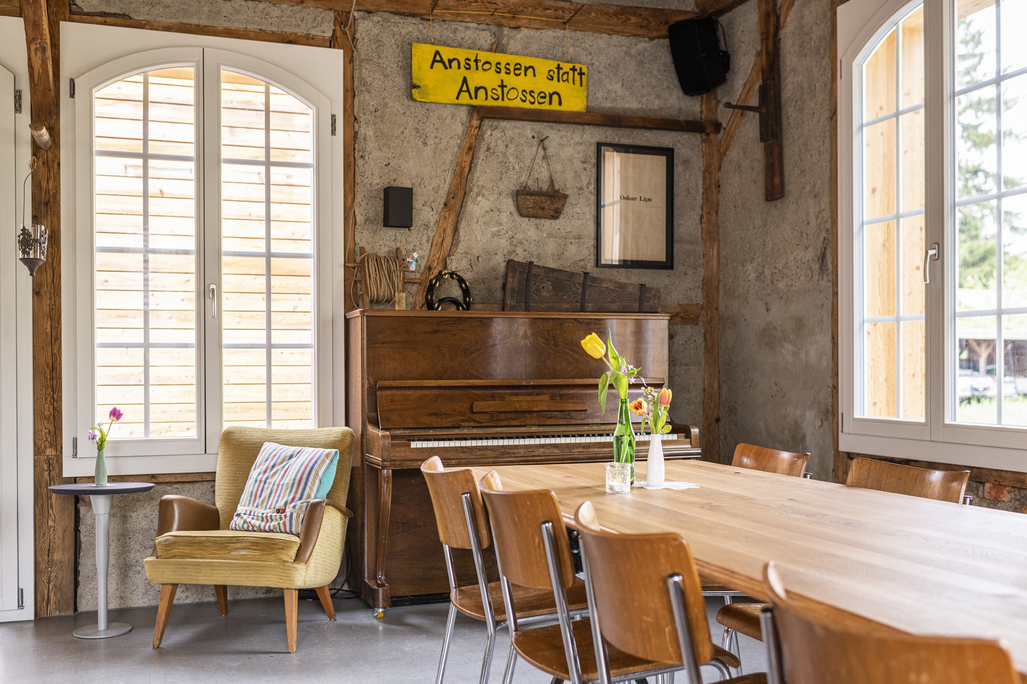 Innenraum der Alten Wagnerei mit Holzdecke, großen Fenstern und einem alten Klavier, auf dem sich Blumenvasen mit Tulpen befinden. Es gibt einen Sitzbereich mit einem bequemen Sessel, einem kleinen Tisch und einer Standleuchte.