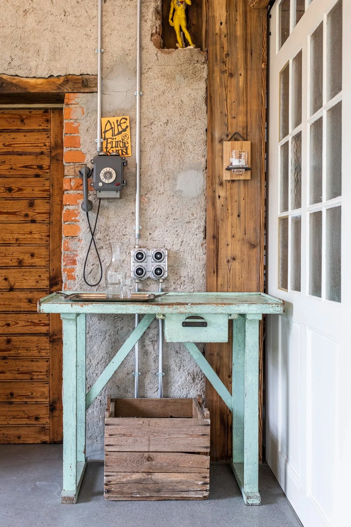 Ein Holztisch vor einer Wand mit Holz und Ziegeln, darauf eine leere Obstkiste, ein altes Wasserfass, eine Telefonzelle und eine kleine Glasvase. Rechts eine Tür mit Glasfenstern.