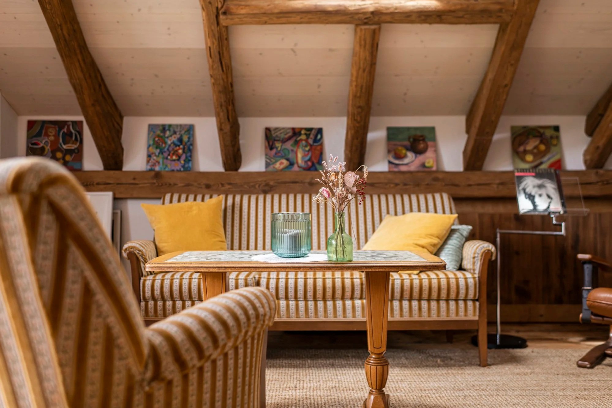 Galerie mit Holzdachbalken, Sessel mit gelben Kissen, Holztisch mit Vase und Blumen, Wandbilder im Hintergrund, Stehlampe auf der rechten Seite.
