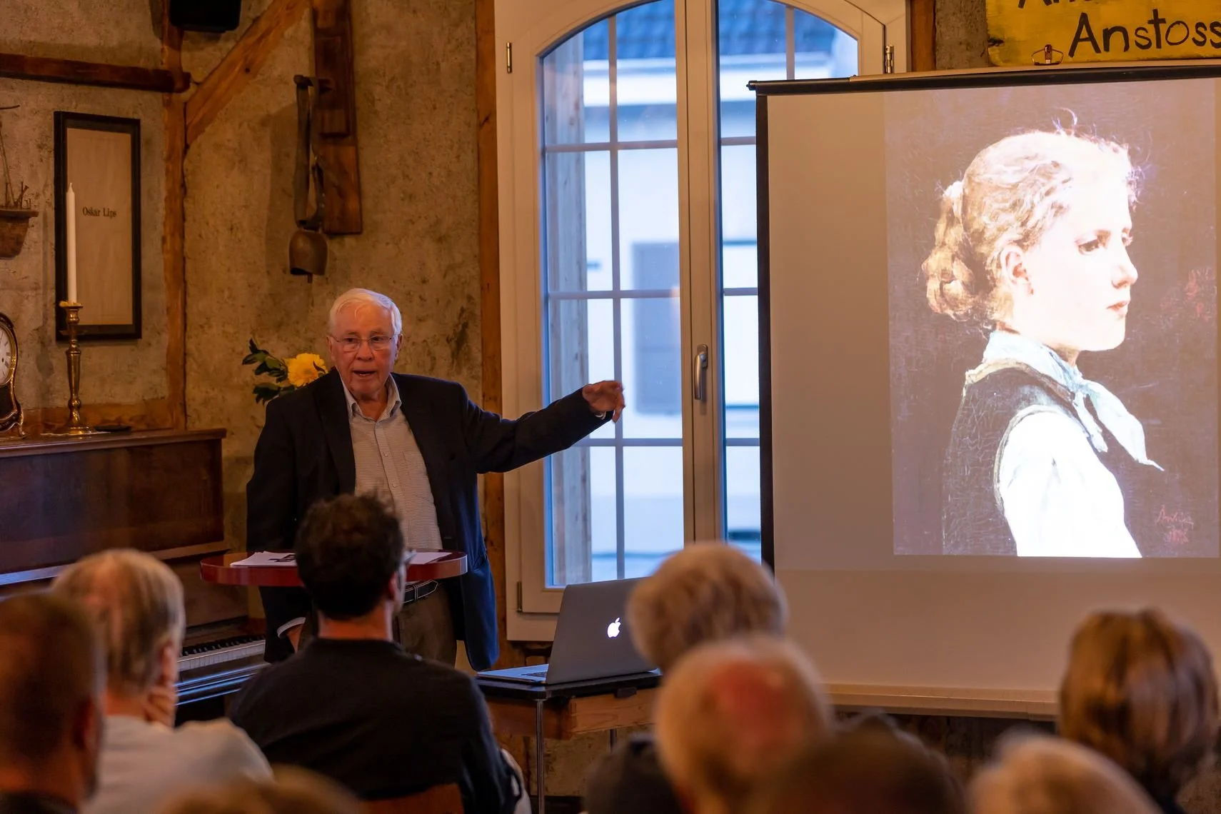 Christoph Blocher hält eine Präsentation vor Publikum über Albert Anker in der Alten Wagnerei. Auf einer Leinwand ist das Profil eines Mädchens zu sehen, das an ein Gemälde erinnert.