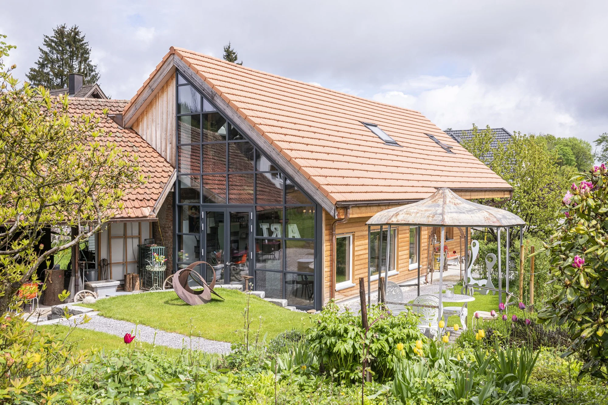 Die Alte Wagnerei mit holzverkleideter Fassade, grossen Fenstern und einem roten Ziegeldach in einem Garten mit bunten Blumen und grünem Rasen.
