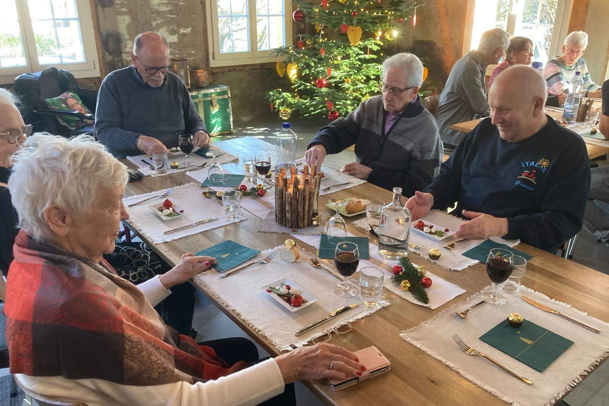 Eine Gruppe älterer Menschen sitzt an einem festlich gedeckten Tisch vor einem Weihnachtsbaum, der mit bunten Ornamenten geschmückt ist, in einer rustikalen Holzhütte, und genießt ein festliches Beisammensein.