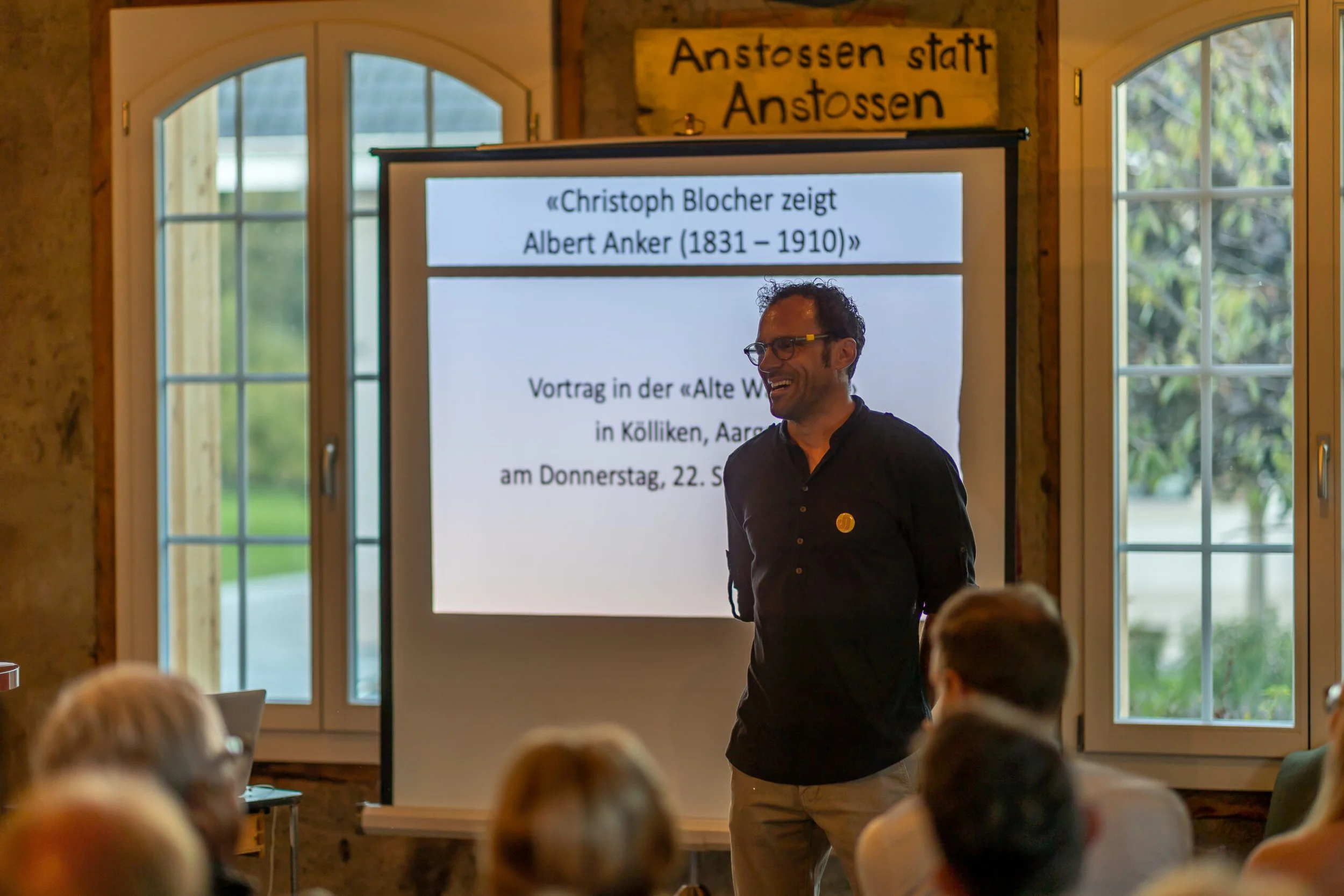 David Spielmann spricht vor Publikum in einem Raum mit grossen Fenstern, eine Präsentationsfolie im Hintergrund. Die Leute im Publikum sind zu sehen, sprechen oder hören zu.