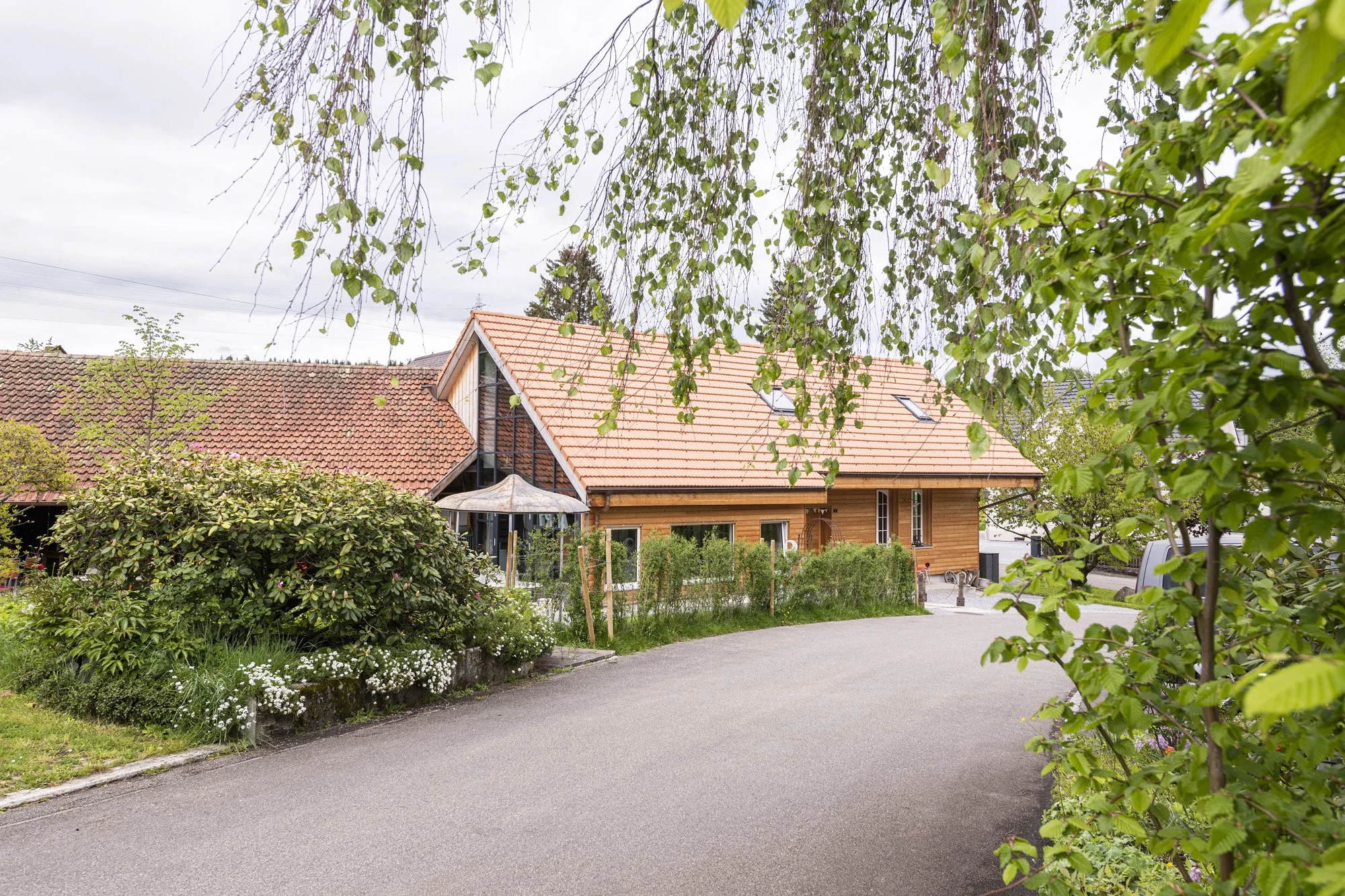 Die Alte Wagnerei mit rotem Ziegeldach, umgeben von einem gepflegten Garten mit Büschen, Blumen und Bäumen, vor einem bewölkten Himmel.