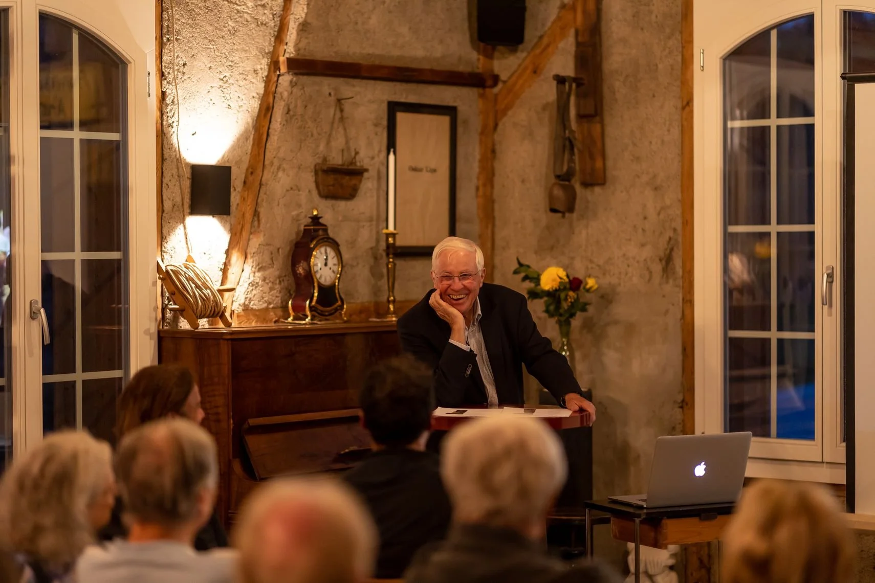 Christoph Blocher lächelt während eines Vortrags in der Alten Wagnerei, umgeben von mehreren Zuhörern, im Vordergrund zu sehen ist eine Gruppe von Menschen, und auf dem Tisch vor ihm liegt ein Laptop.