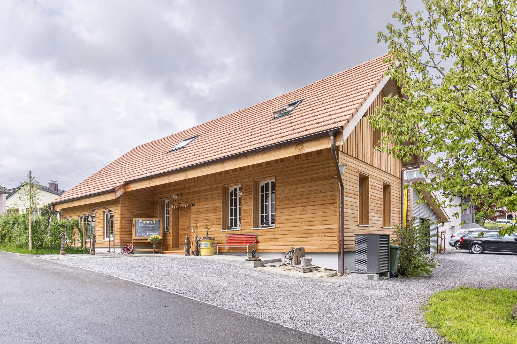 Die Alte Wagnerei mit rotem Ziegeldach, grenzt an eine Strasse, umgeben von Grünflächen und Bäumen, bei bewölktem Himmel.