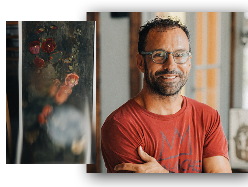 David Spielmann, lachend, in einem roten T-Shirt steht vor einem Gemälde, das Blumen zeigt, in einem Raum mit großen Fenstern im Hintergrund.