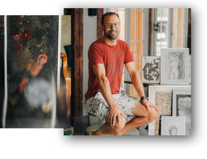 David Spielmann mit Brille, rotem T-Shirt, Shorts und Barfuss sitzt in der Alten Wagnerei neben Gemälden und Kunstwerken.