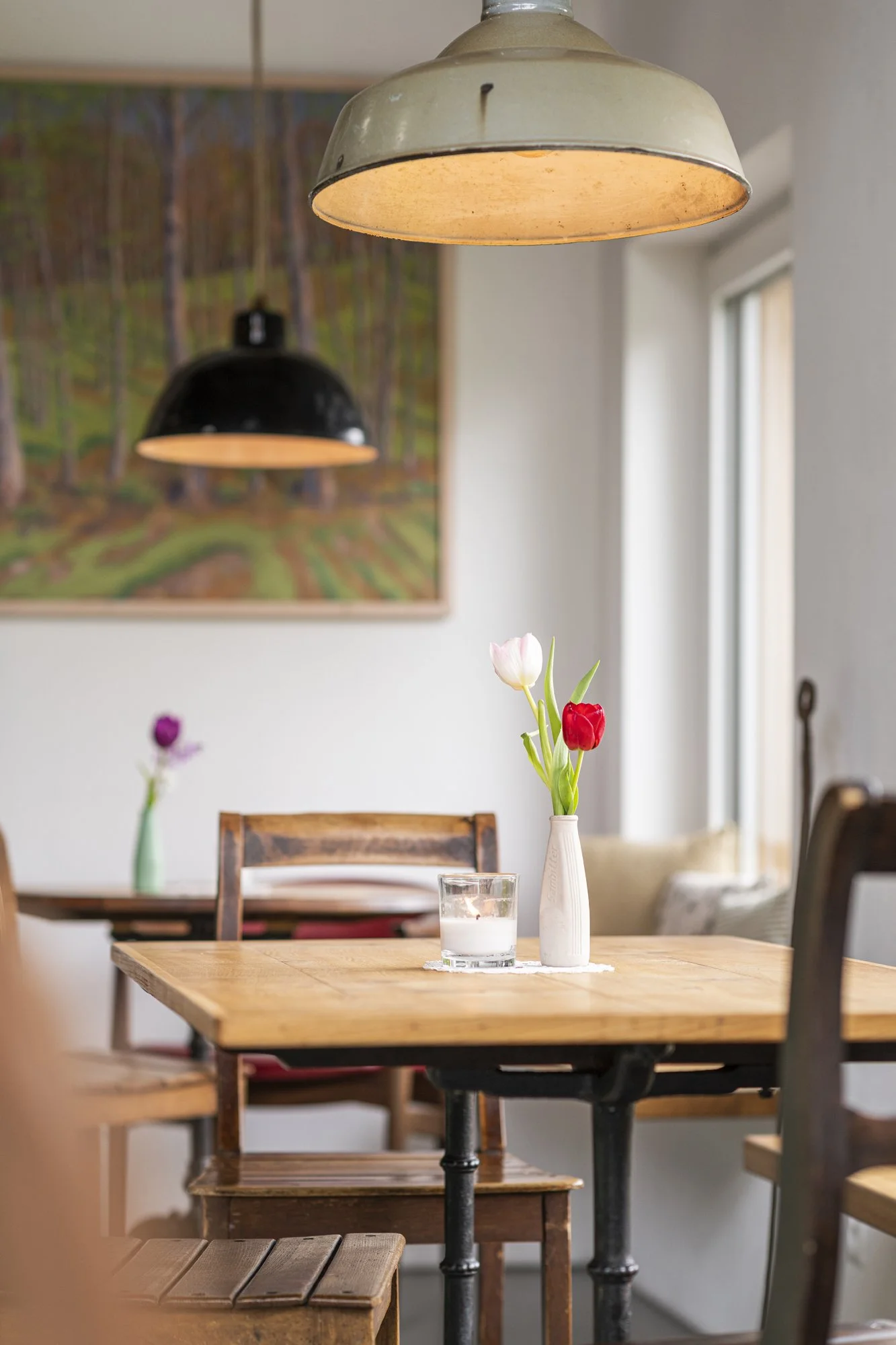 Ein Holztisch mit einer weissen Vase und Tulpen sowie einem Glas Wasser in einem hellen Raum, mit Hintergrundbildern an der Wand und einer Stehlampe.