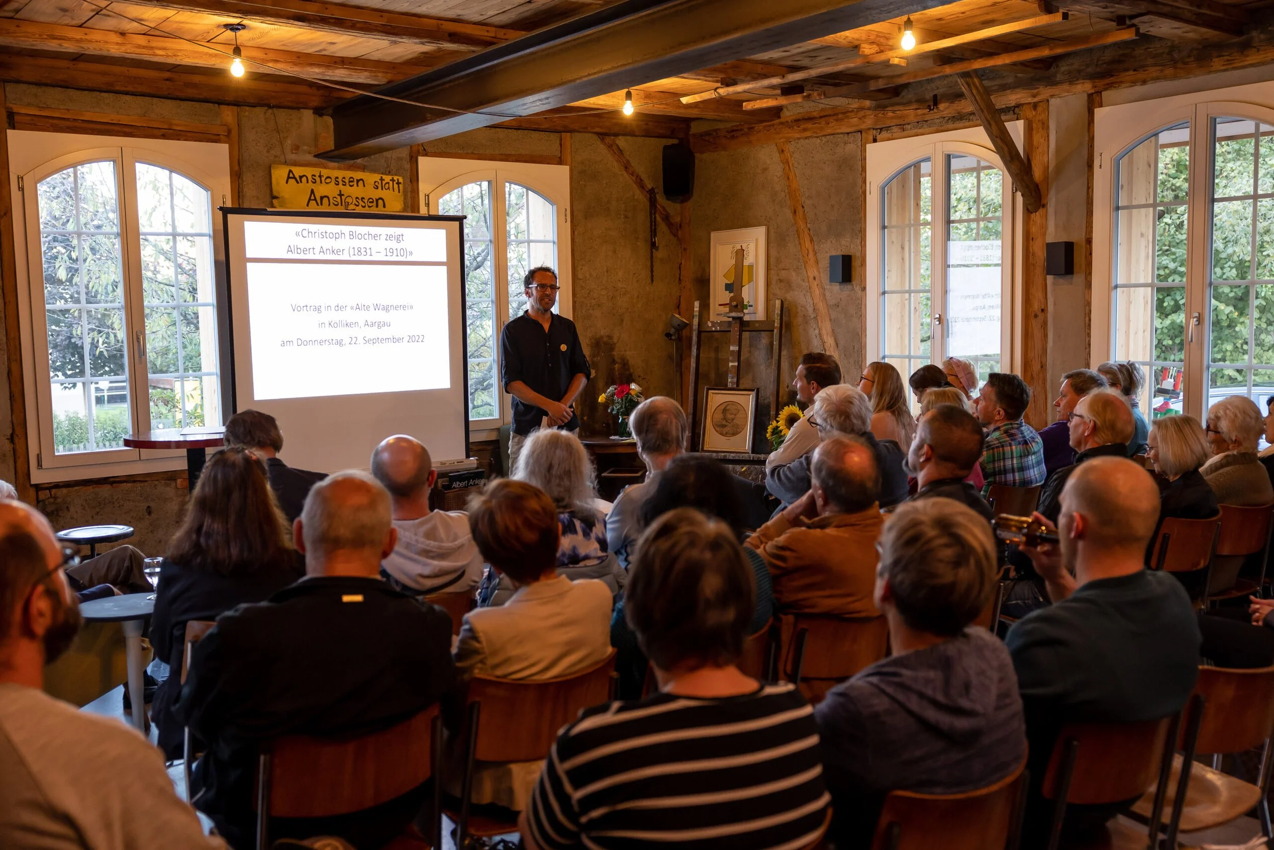 Ein Vortrag wird in einem rustikal gestalteten Raum gehalten, mit Holzdecke und großen Fenstern. Ein Mann steht vor einer Leinwand, auf der Text angezeigt wird. Das Publikum sitzt und hört zu.