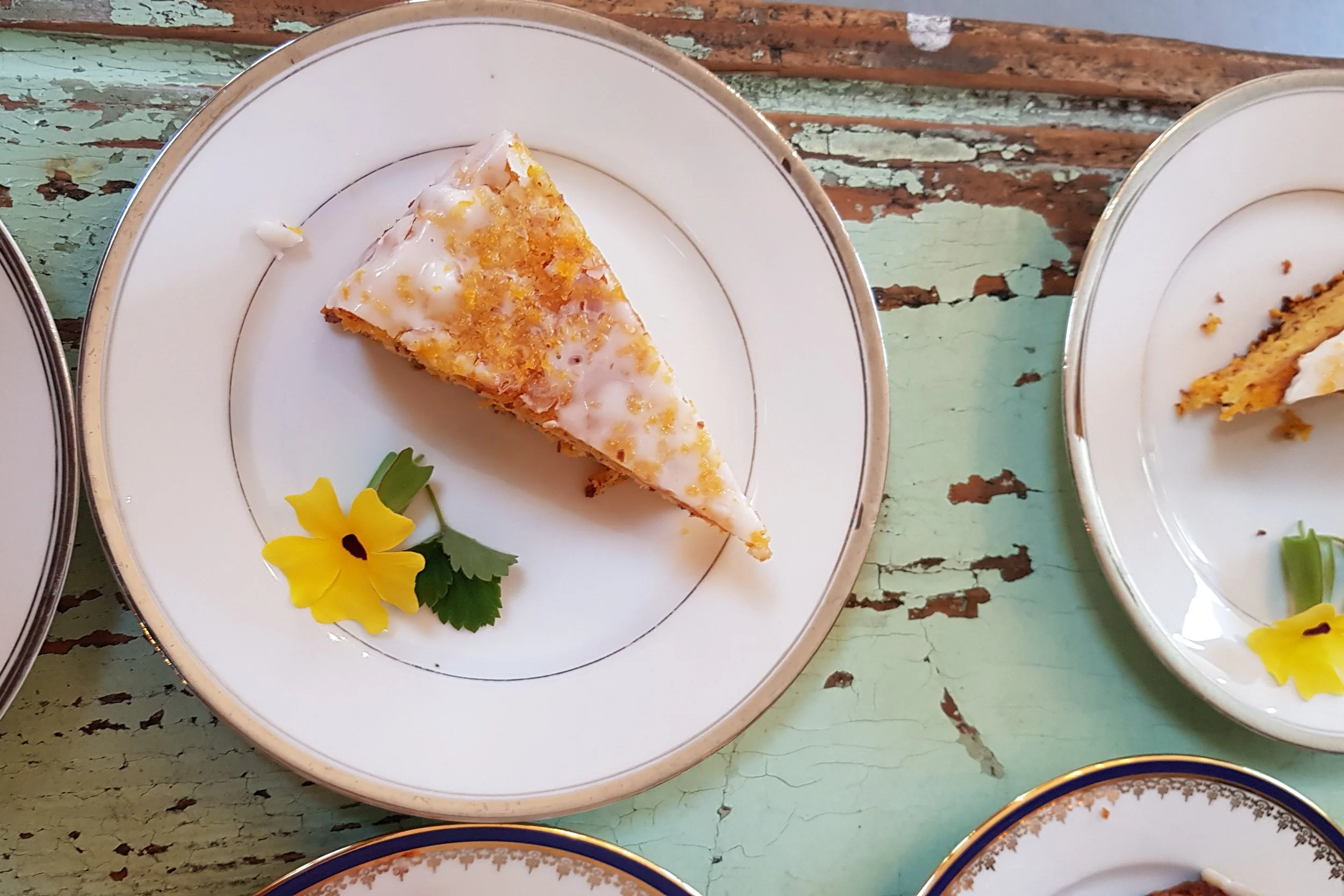 Ein Stück Kuchen mit weißer Glasur und Zitruszesten auf einem weißen Teller, verziert mit einer gelben Blume und grüner Petersilie auf einem alten, grünlichen Holz-Tisch.