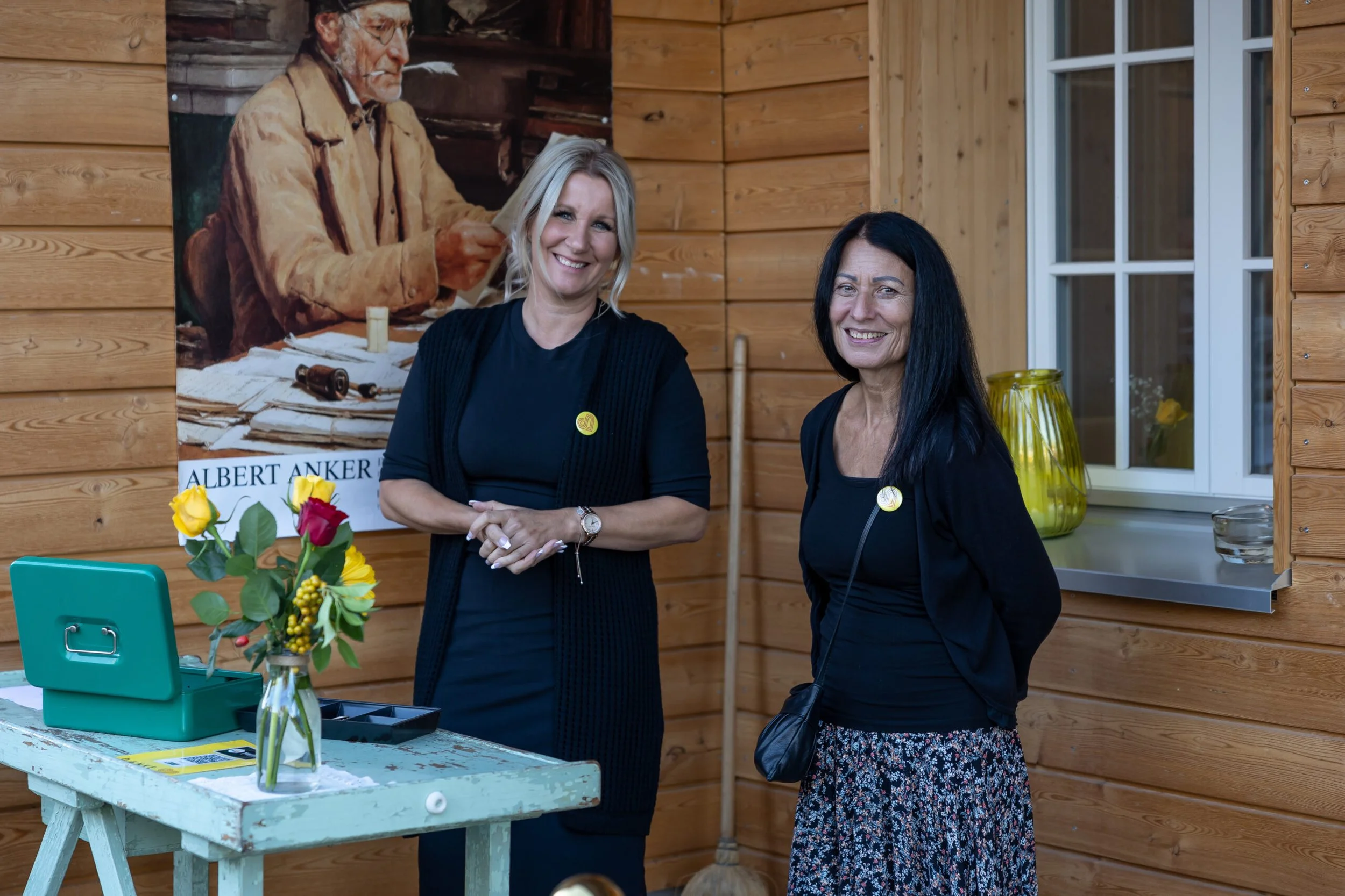 Zwei Frauen stehen in einem Raum mit Holzverkleidung, lächeln zur Kamera, im Hintergrund ein Poster. Vor ihnen ein alter, verzauberter Tisch mit bunten Blumen in einer Vase und einem grünen Kasten. Sie tragen schwarze Kleidung, die eine Frau hat lang