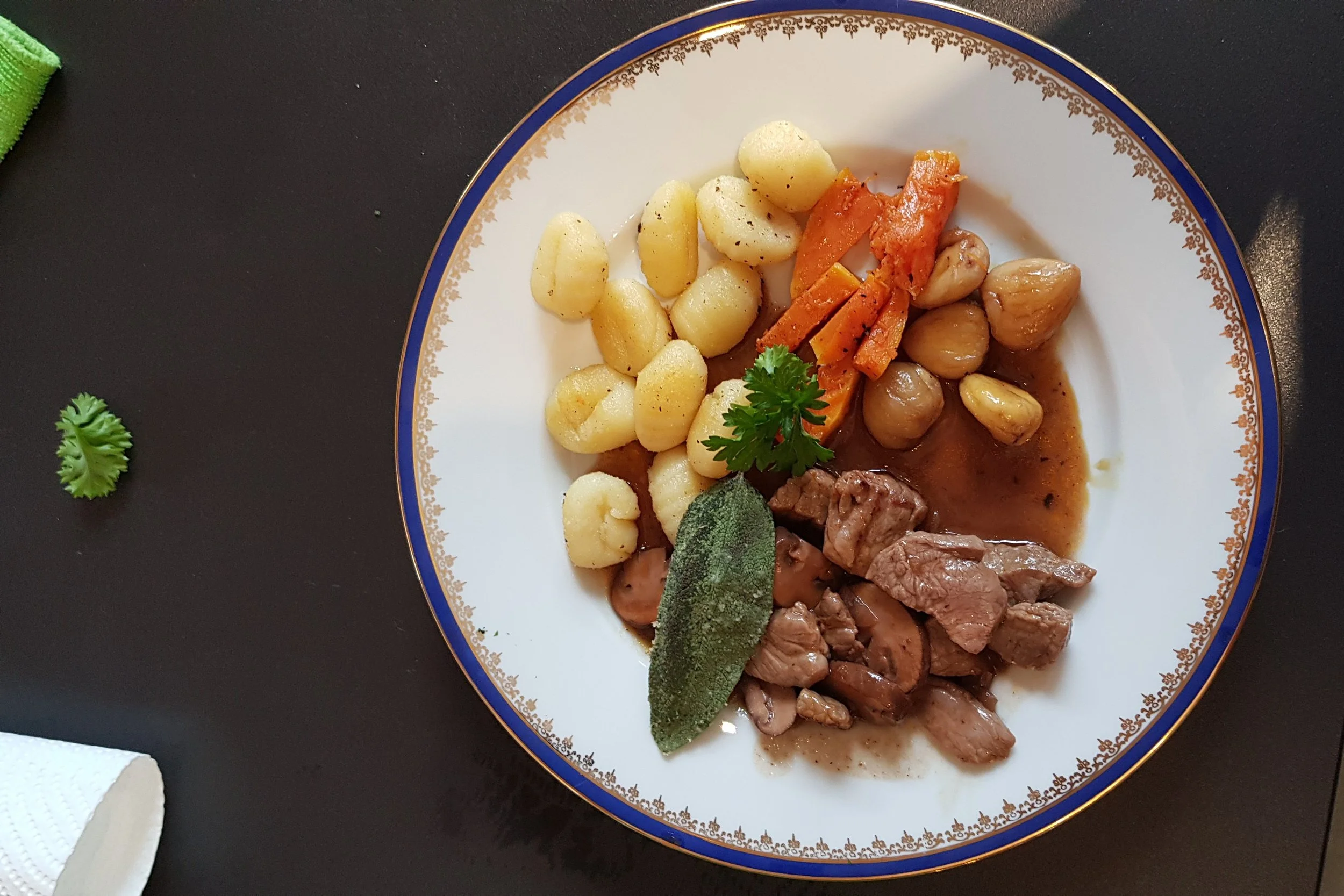 Ein Teller mit Gnocchi, Karotten, Pilzen, Rindfleisch, Salbei und einer braunen Soße auf einem schwarzen Tisch.