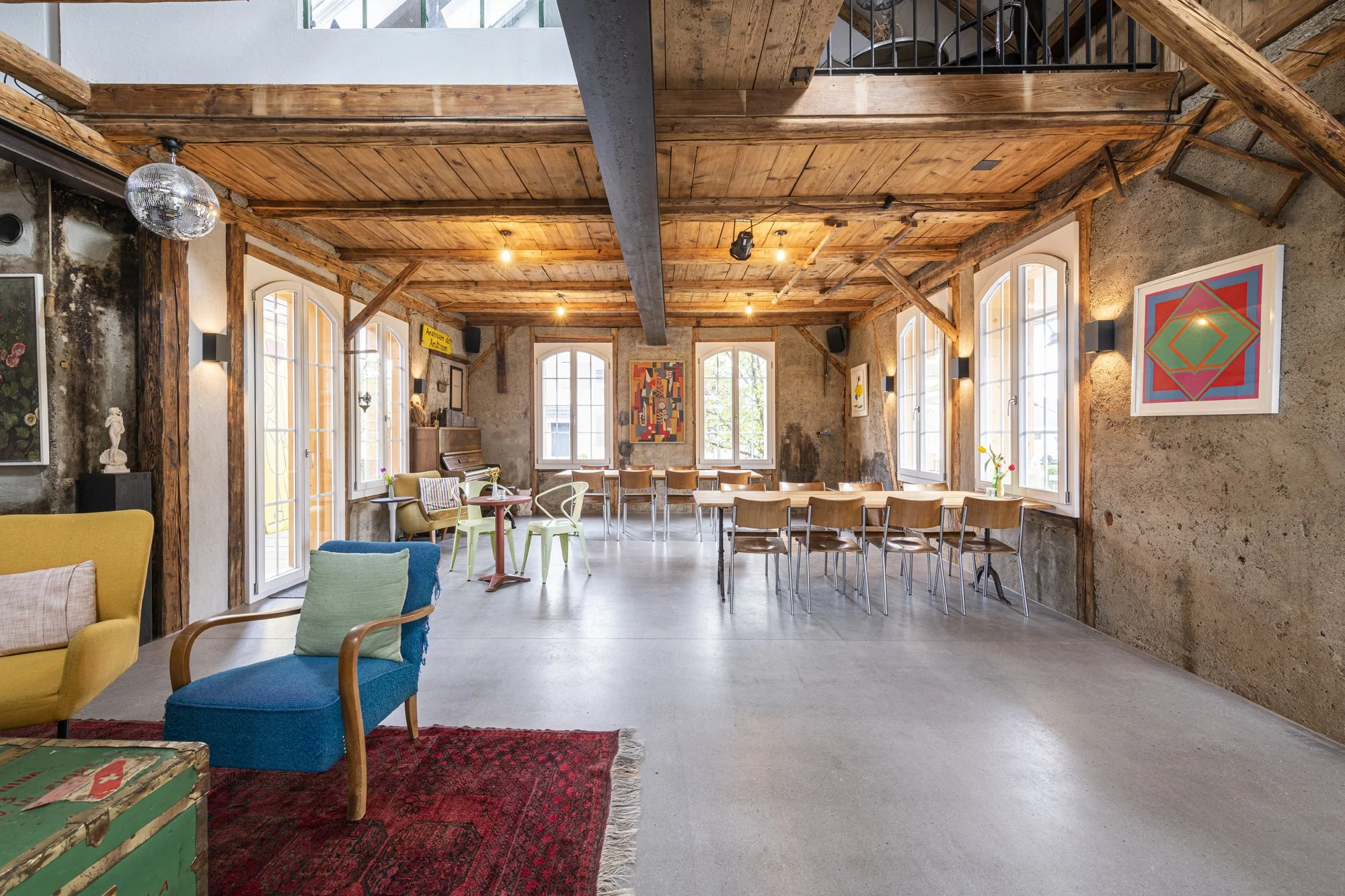 Innenraum der alten Wagnerei mit Holzbalken und Kunstwerken an den Wänden, mehreren Stühlen, einem Klavier, einem Disco-Kugel und einem verschiedenen Sitzmöbeln.