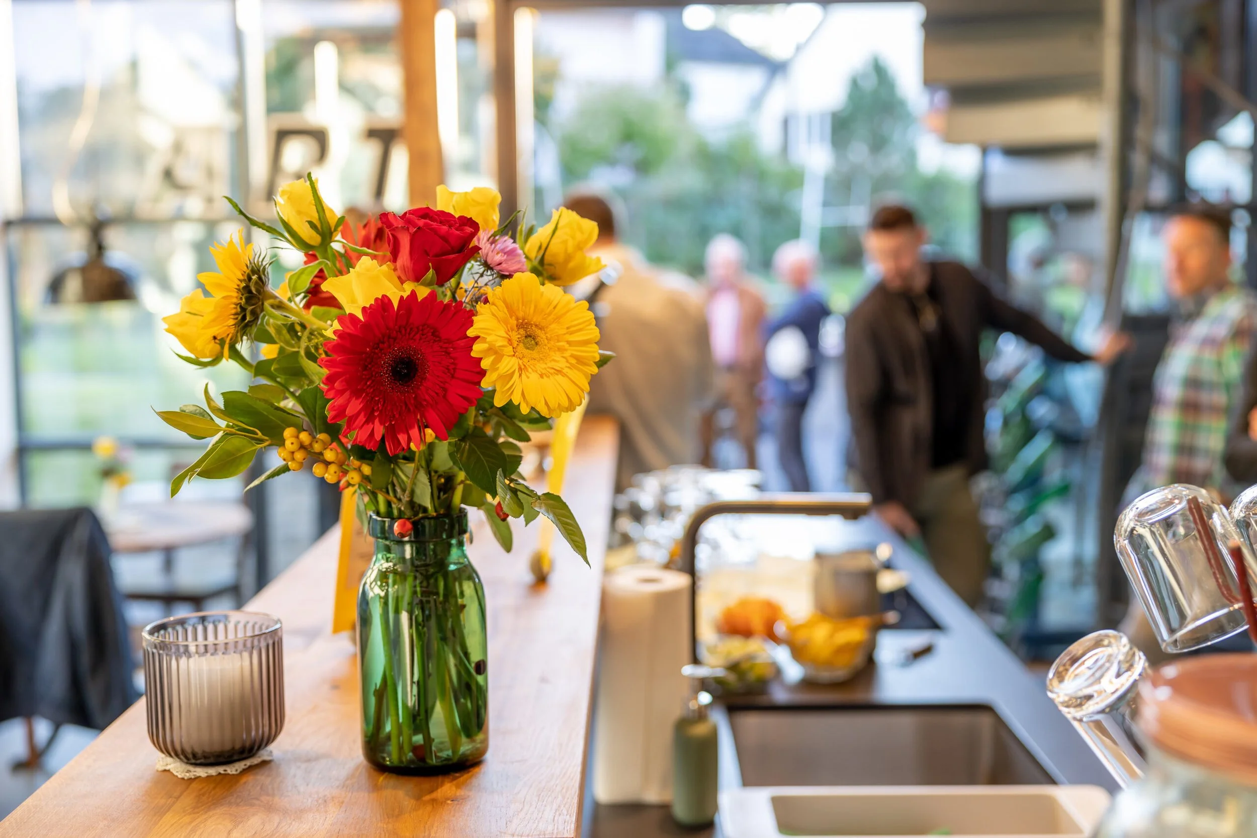 Blumenstrauss mit roten, gelben, pinken Blumen in einer grünen Glasvase auf einer Holztisch im Hintergrund Menschen in einer modernen Küche oder Cafeteria