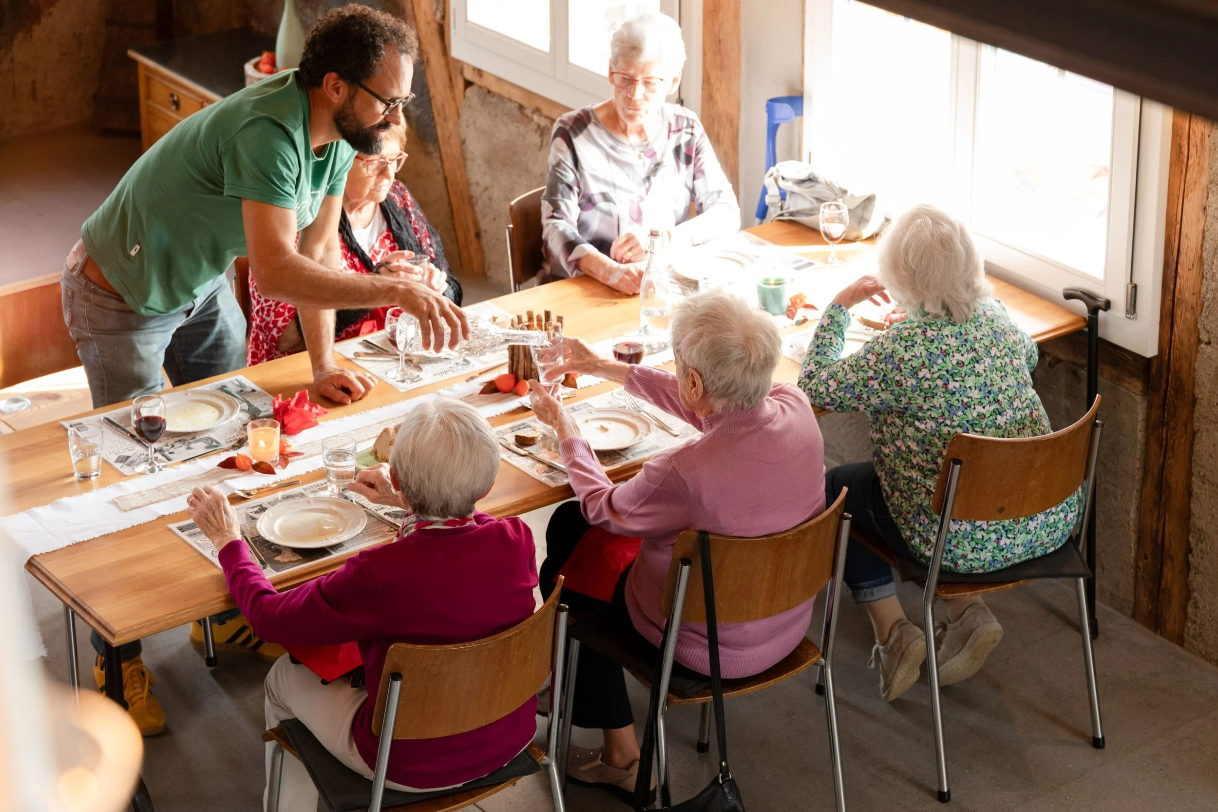 Eine Gruppe älterer Menschen sitzt und steht an einem Esstisch, der in einem rustikalen Raum mit Holzwänden steht. Sie essen, trinken und unterhalten sich. Es ist tagsüber, das Licht fällt durch Fenster auf den Tisch.