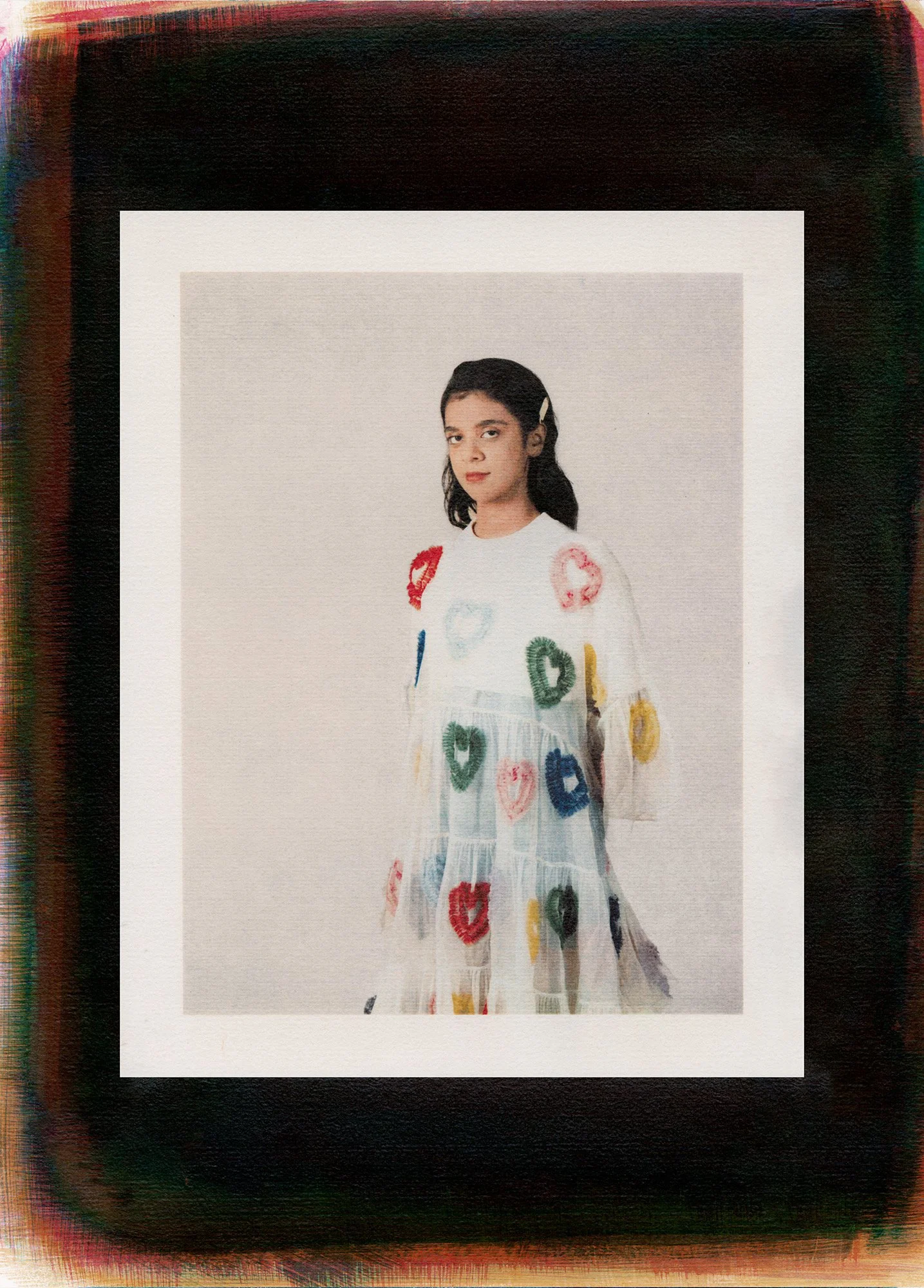 A young woman with dark hair wearing a white dress decorated with colorful hearts.