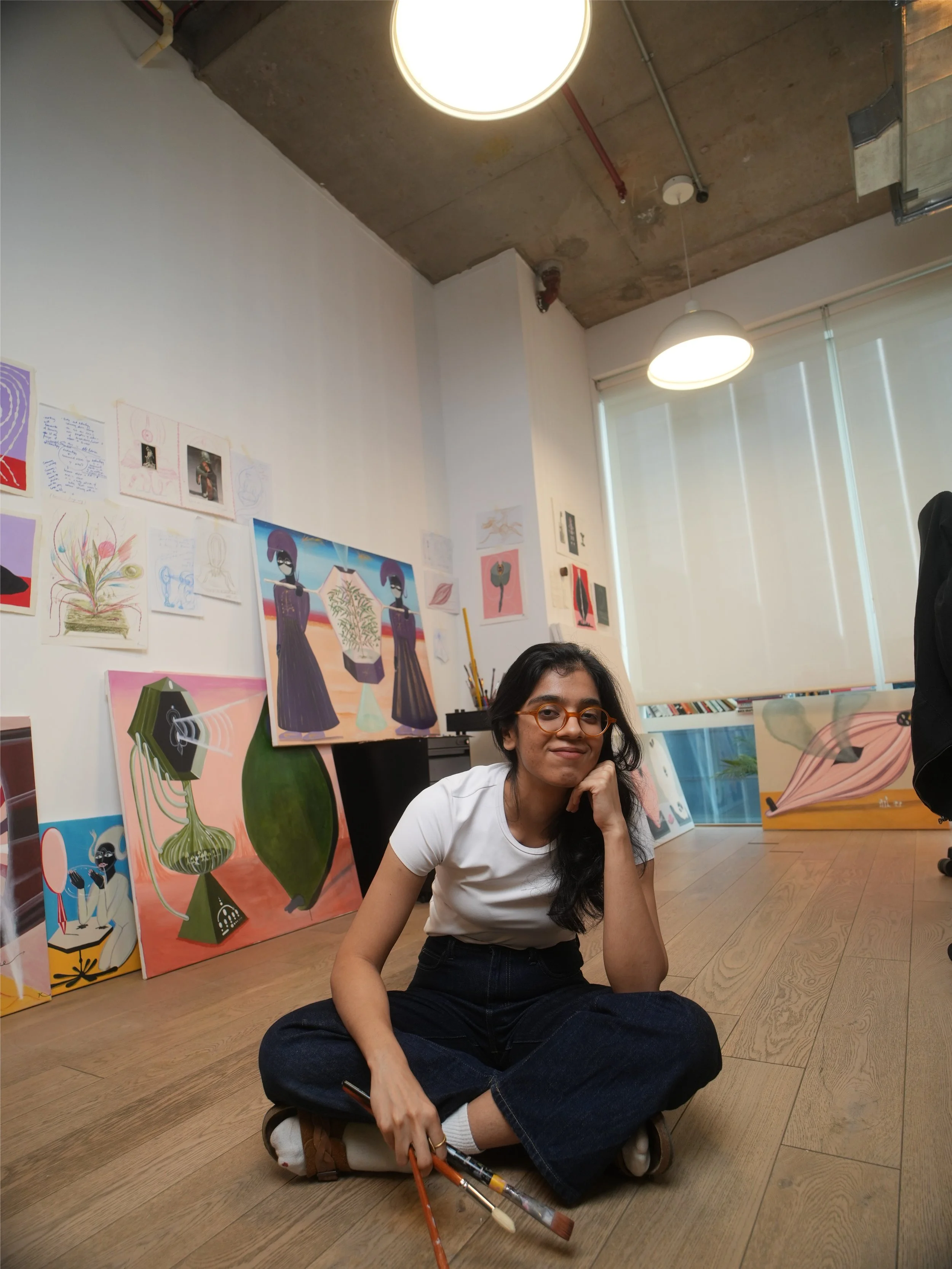 A woman with glasses sitting cross-legged on her art studio floor, smiling at the camera. Behind her are colorful paintings of abstract figures and plants displayed on the wall, with large window blinds partially open letting in natural light.