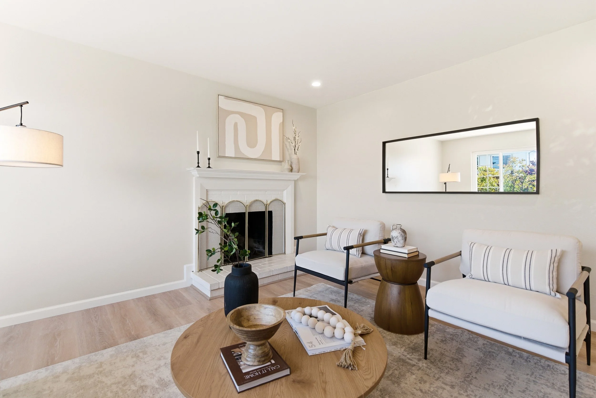 Living room with a white fireplace, two chairs with striped cushions, a wooden side table, a large wall mirror, and window showing trees outside.