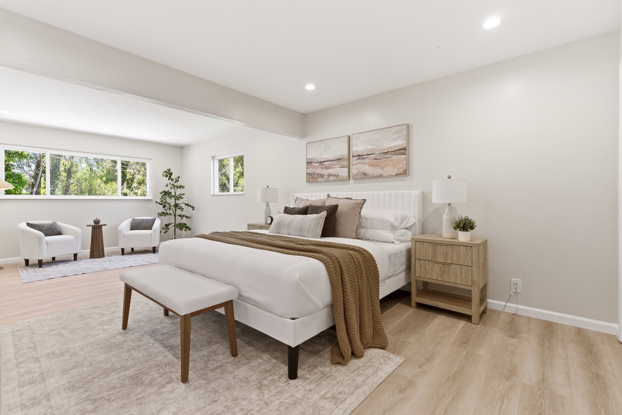 Bright bedroom with white walls, large window with greenery outside, a bed with neutral bedding and pillows, two white armchairs, a small wooden table, a large potted plant, wall art, and a nightstand with a lamp and decor items.