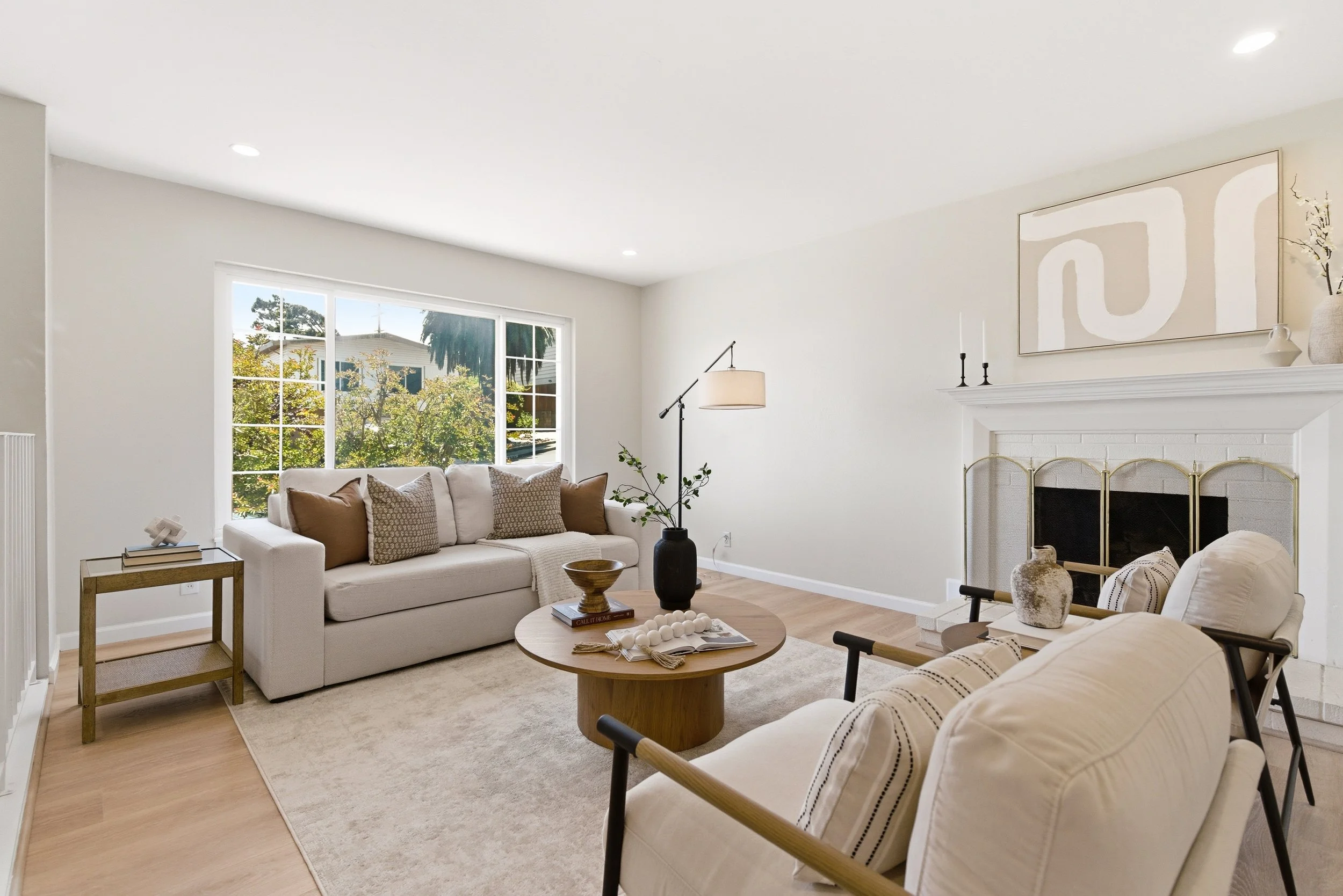 Bright living room with large window, white walls, and minimalist decor including a white sofa with patterned cushions, a round wooden coffee table with books and decorative objects, and a fireplace with a mantel decorated with candles and vases.