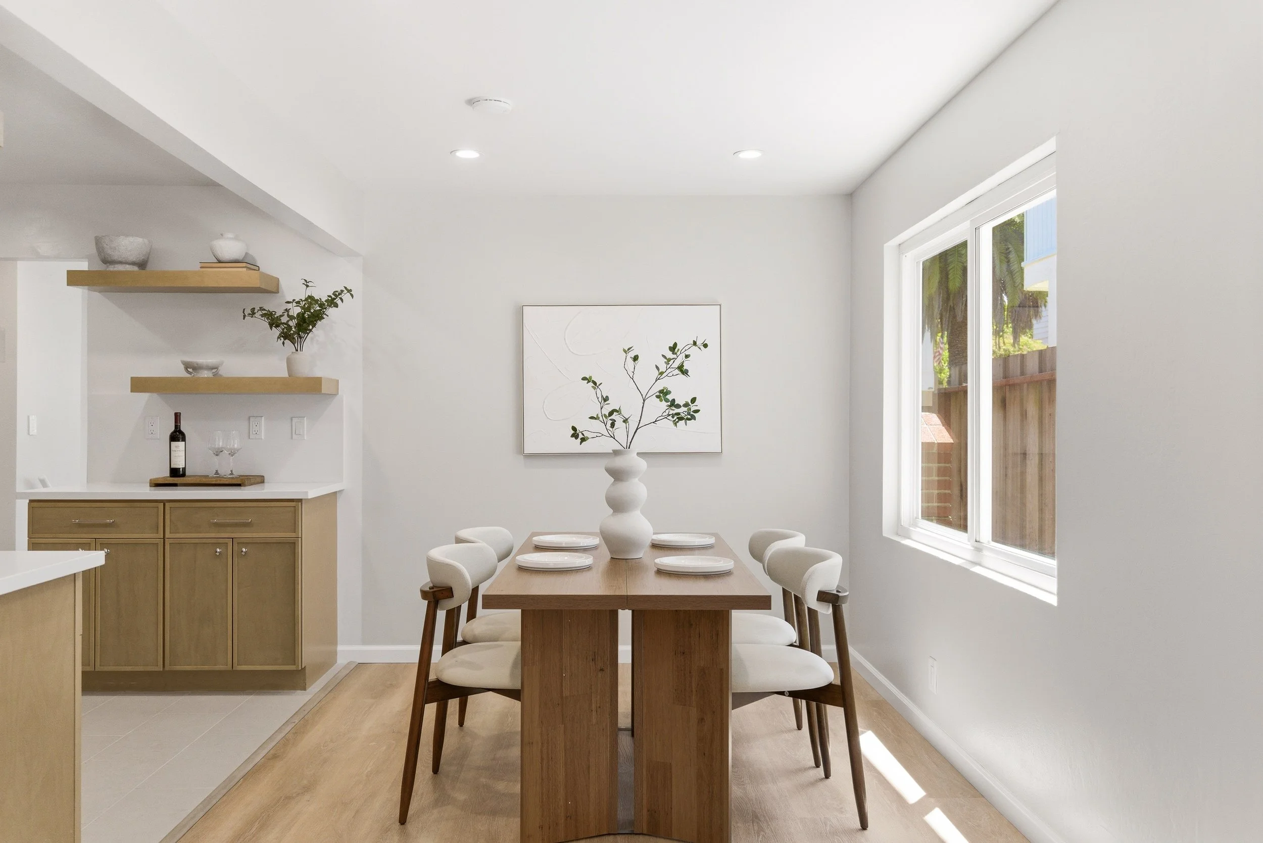 Dining area with wooden table and white chairs, large window with view outside, minimalistic decor, and light-colored flooring.
