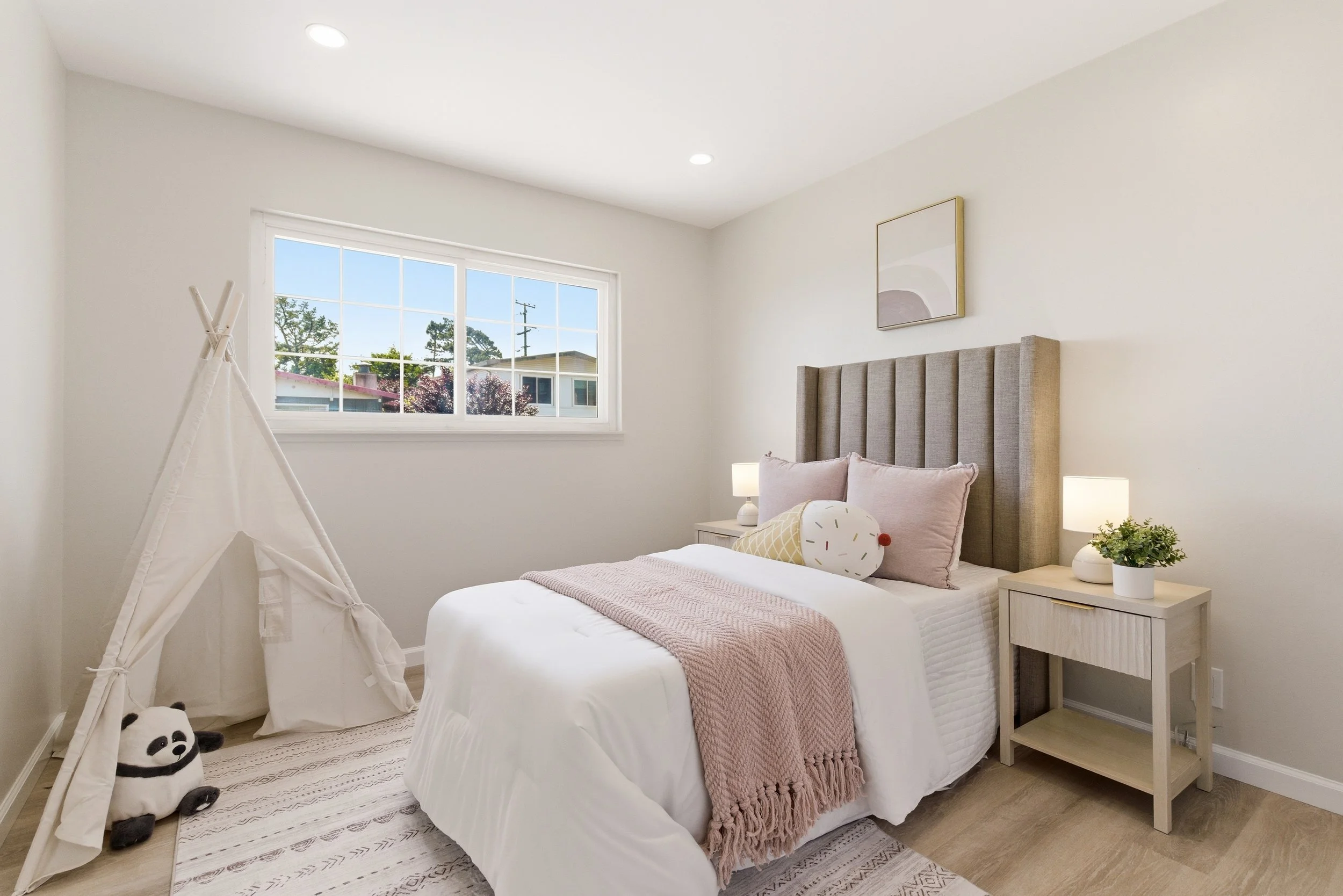 Bright bedroom with white walls, a bed with light pink blankets and pillows, a nightstand with a lamp and plant, a window showing trees outside, a framed minimalist art piece, a white teepee with a panda plush toy inside, and light-colored hardwood f
