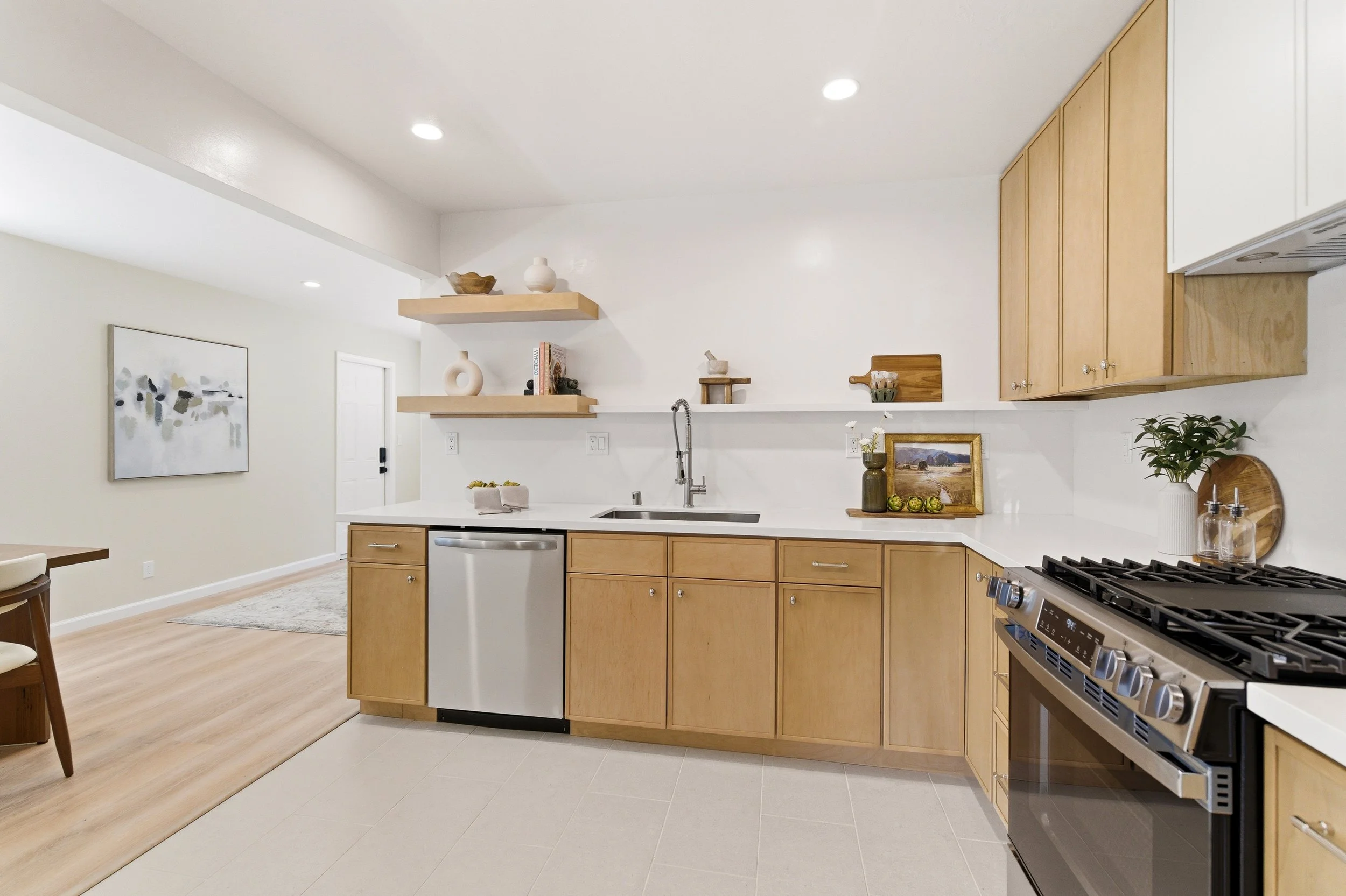 A modern kitchen with light wood cabinets, a white countertop, a stainless steel stove, and minimal decor including framed artwork, vases, and plants.