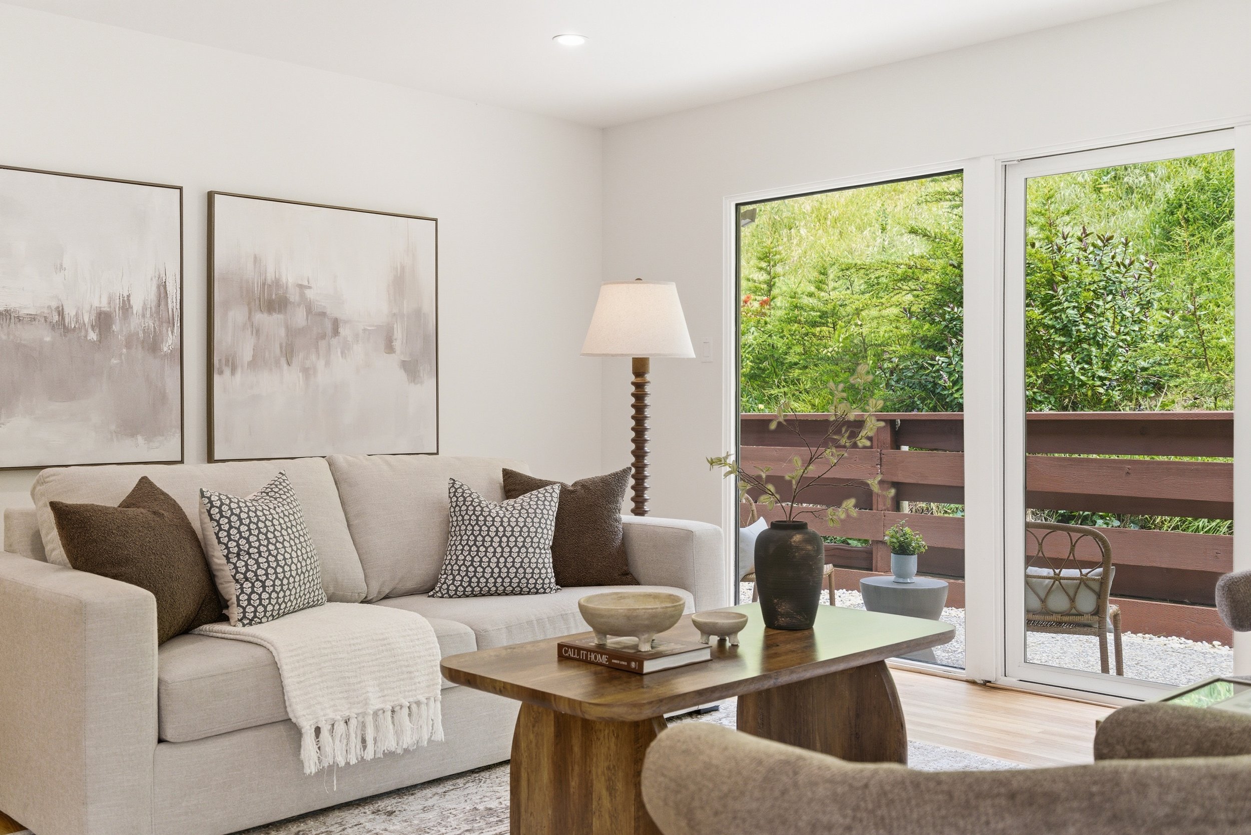 Bright living room with white sofa and patterned pillows, wooden coffee table with decor, large windows showing greenery, and white walls with abstract artwork.