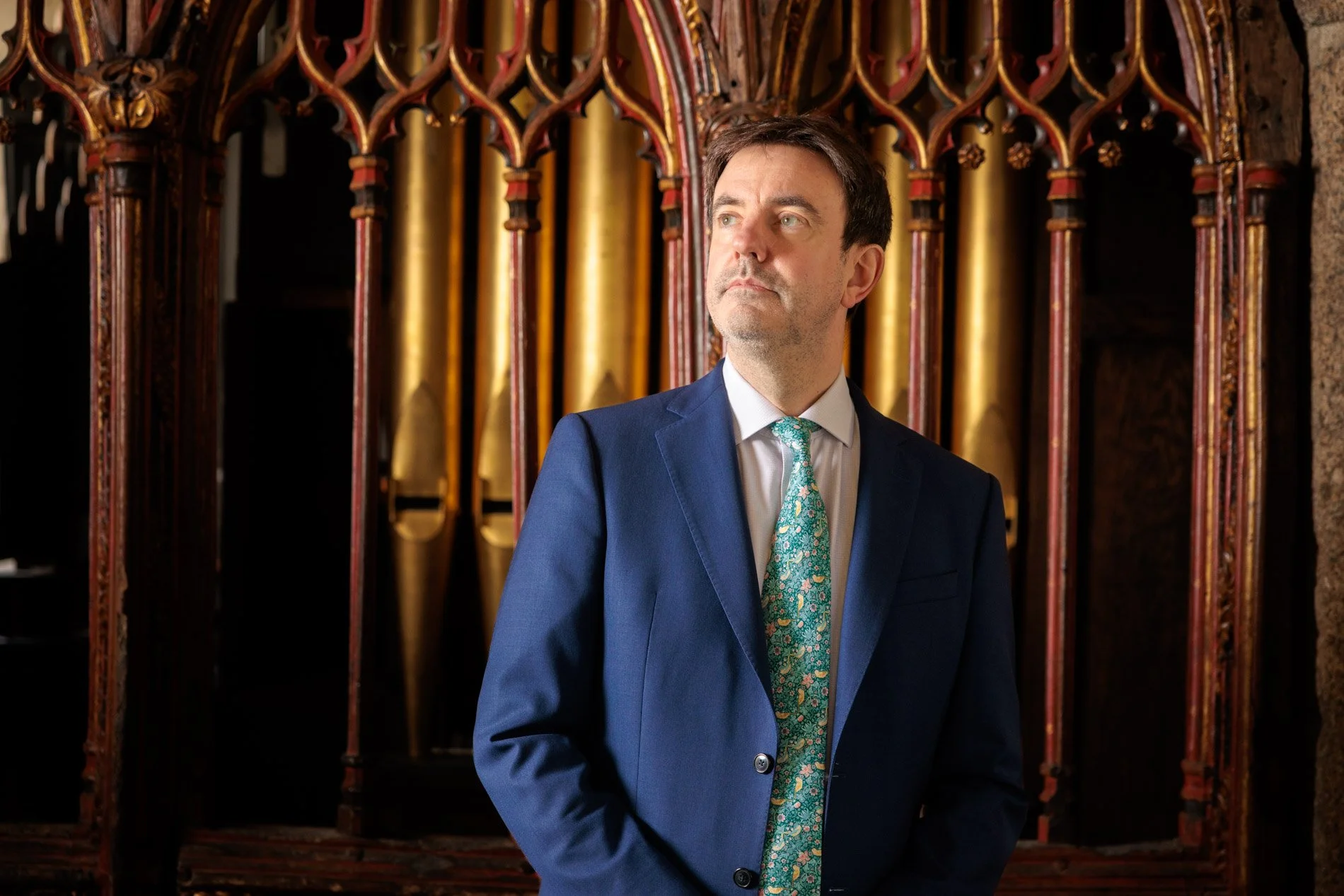 A man in a blue suit with a green patterned tie stands in front of old-fashioned pipe organs with wooden and gold details.