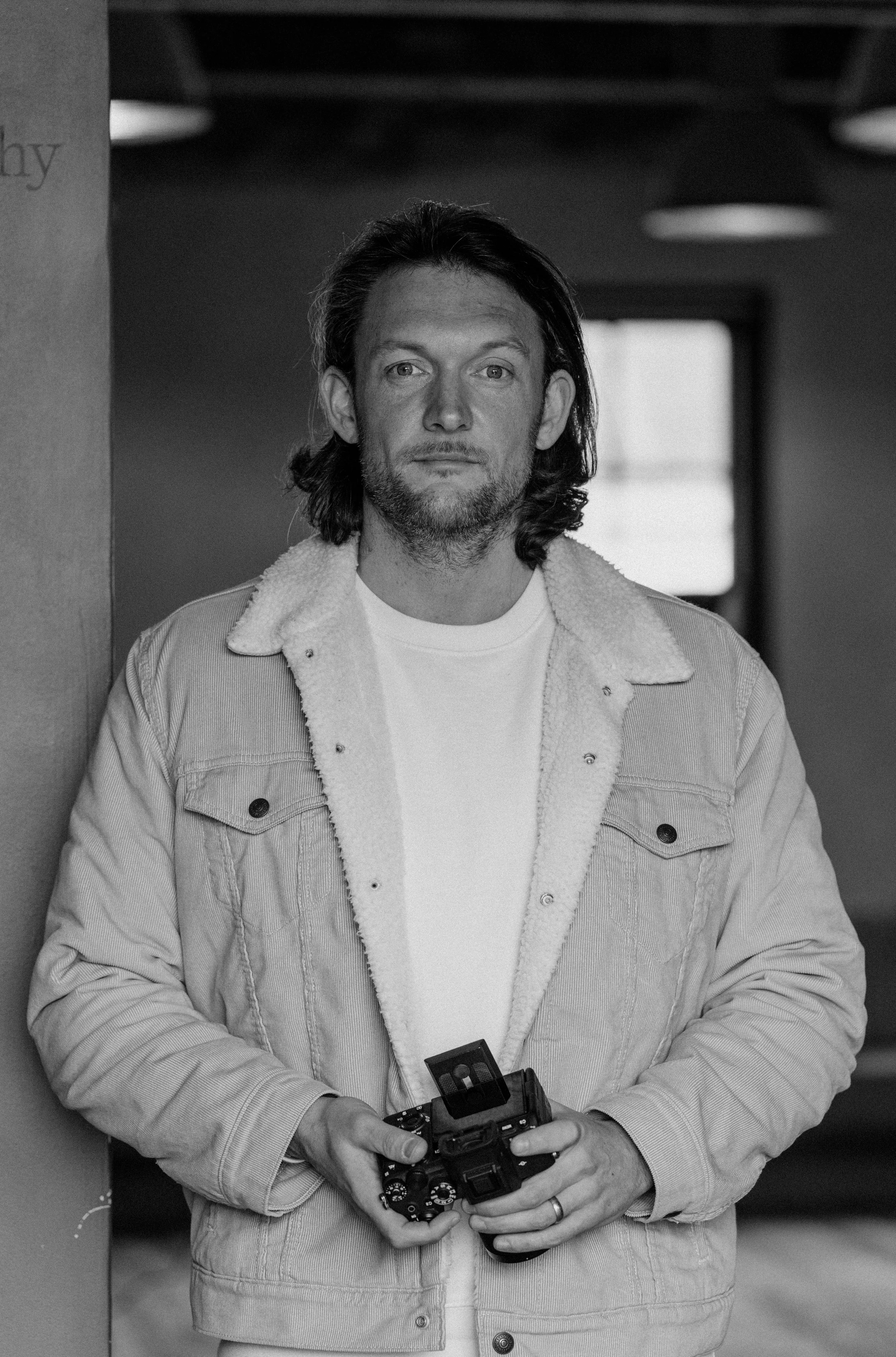 A man with long hair and a beard wearing a light-colored jacket with a fleece collar, holding a camera, standing indoors.