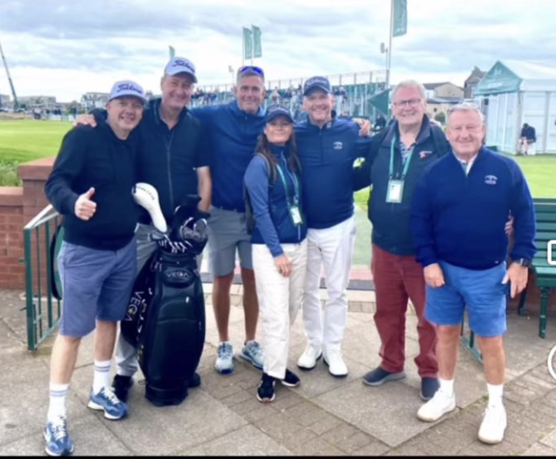 A group of seven people on a golf course, smiling for a photo. Six are standing with arms around each other, one in front wearing a golf glove. They are dressed in golf attire, some wearing caps. Background shows grass, tents, and a cloudy sky.
