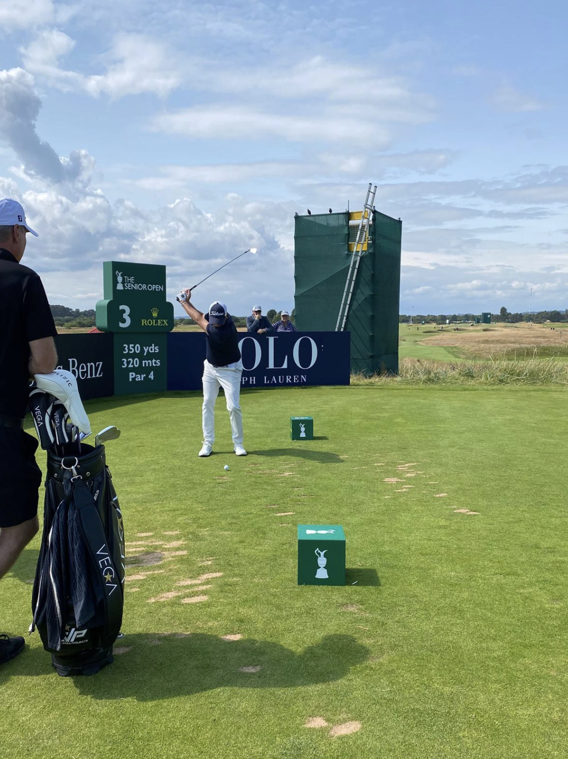 A golfer in white pants and a navy shirt swinging a golf club at the tee box during a tournament at the Senior Open, with a caddy watching, lounge chairs in the background, and large advertisements on the sidelines.