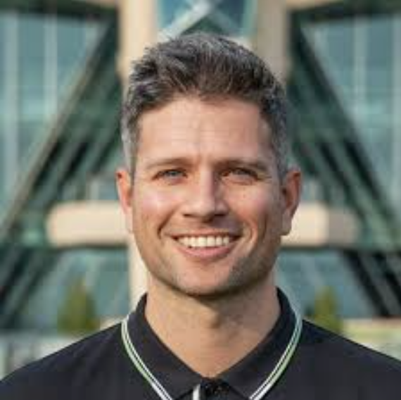 Portrait of a smiling man with short dark hair, wearing a dark collared shirt with a light-colored trim, standing in front of a modern building with glass and metal architecture.