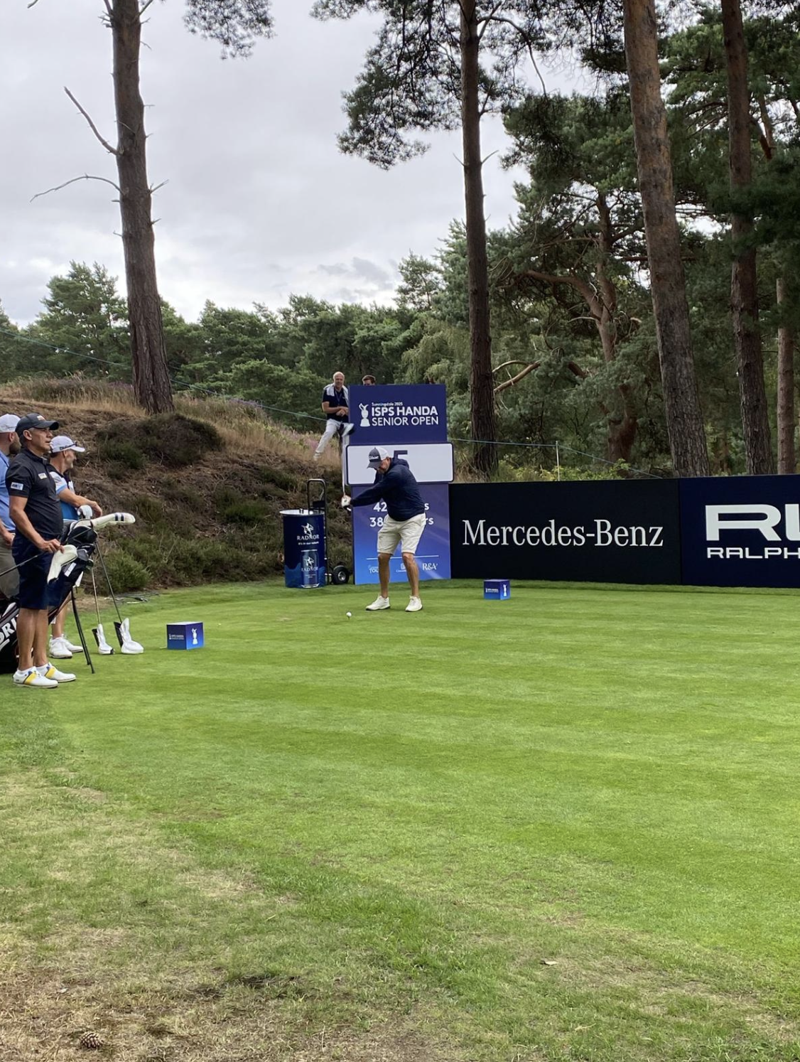 A golfer preparing to hit a shot on a golf course surrounded by trees, with caddies and spectators nearby, during a tournament with signs and banners from sponsors.
