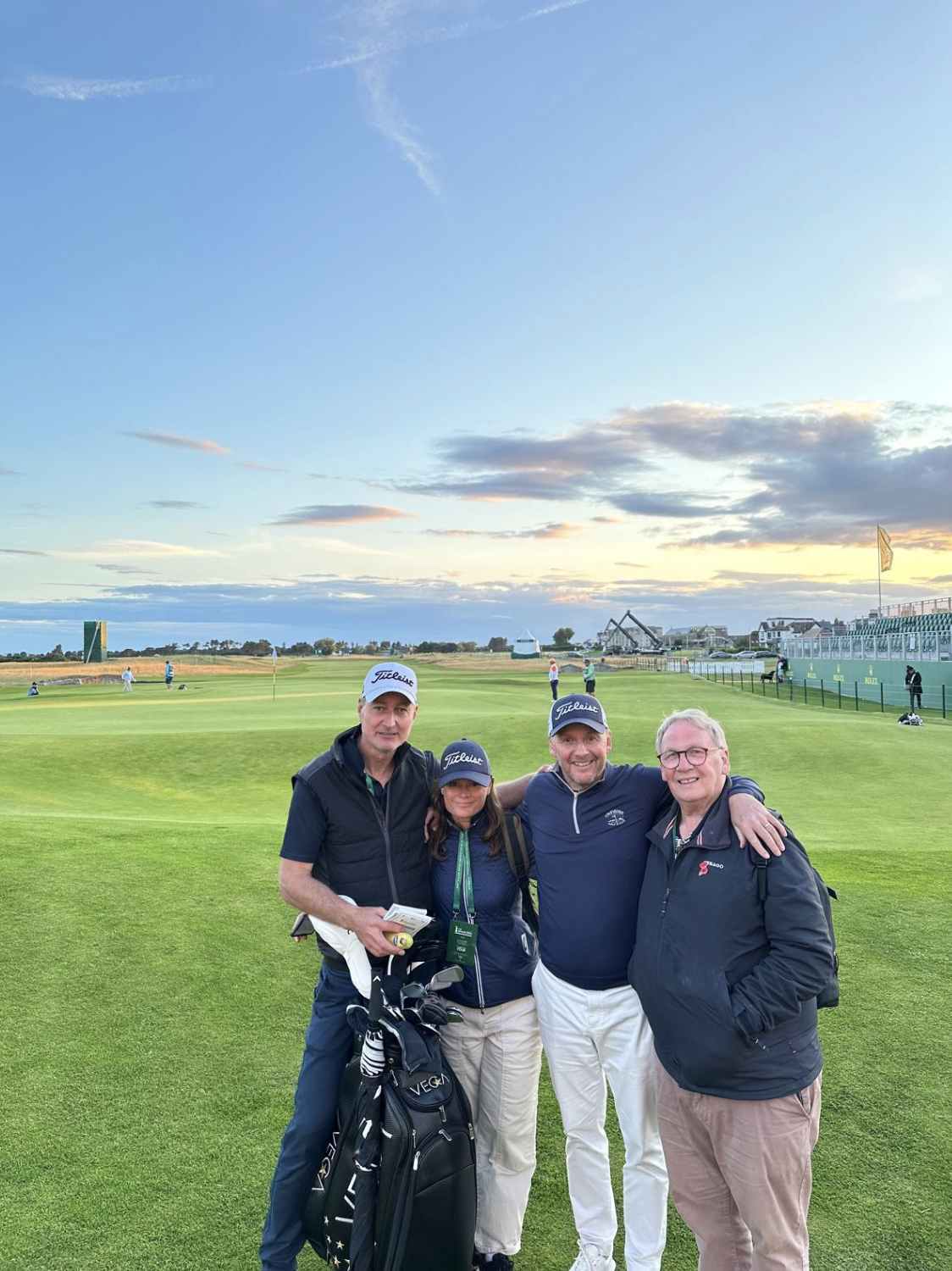 Four people standing on a golf course at sunset, smiling, wearing golf caps and casual golf attire, with golf clubs and a golf bag.