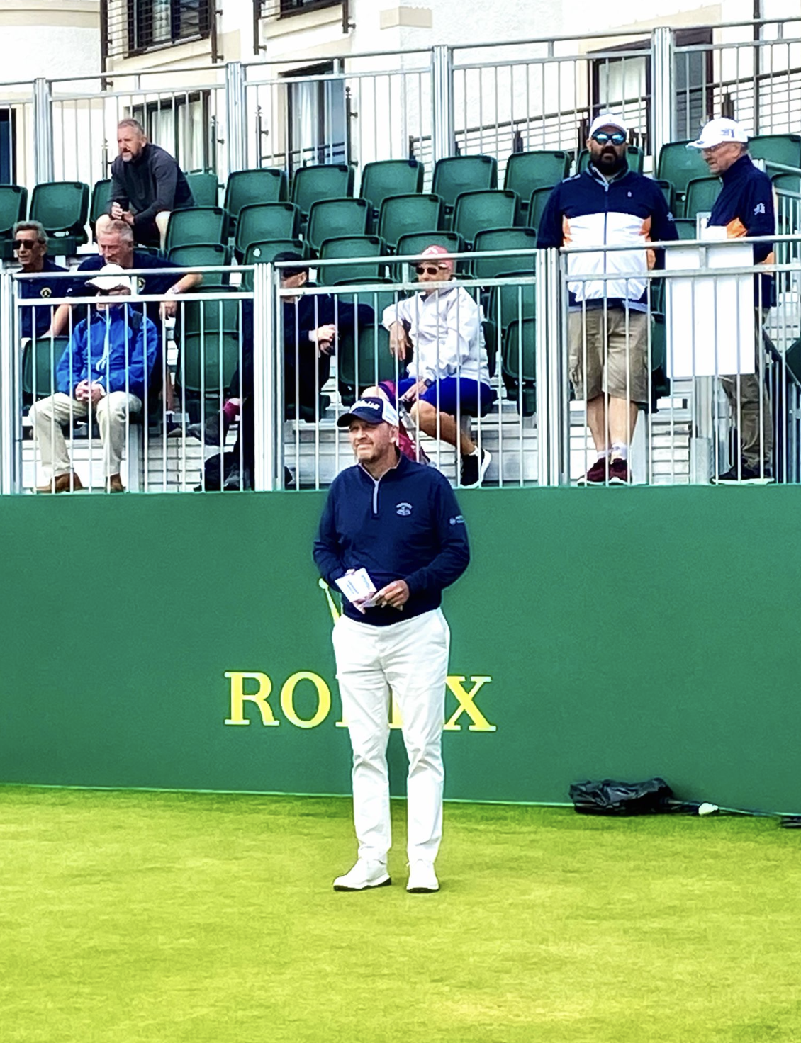A man dressed in white pants and a dark jacket standing on a green sports field holding notebooks, with a green and yellow 'ROLEX' sign behind him. Several people are seated and standing in the background, some watching and some engaged in conversation.