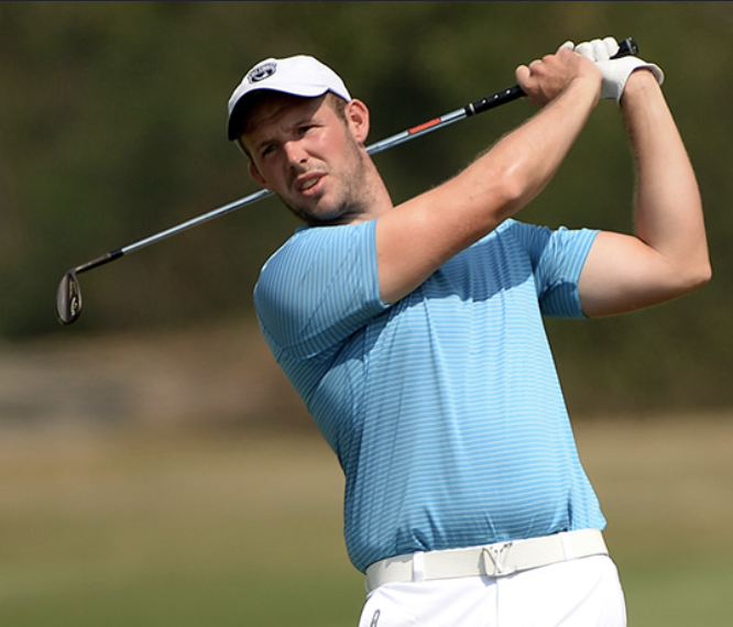 Male golfer in blue shirt and white cap swinging a golf club on a golf course.