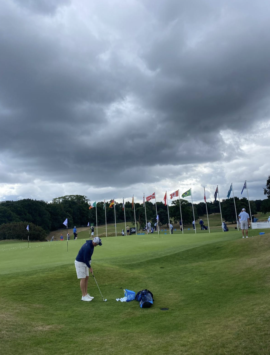 Golf practice range with a golfer preparing to swing, surrounded by flags from various countries, under a cloudy sky.